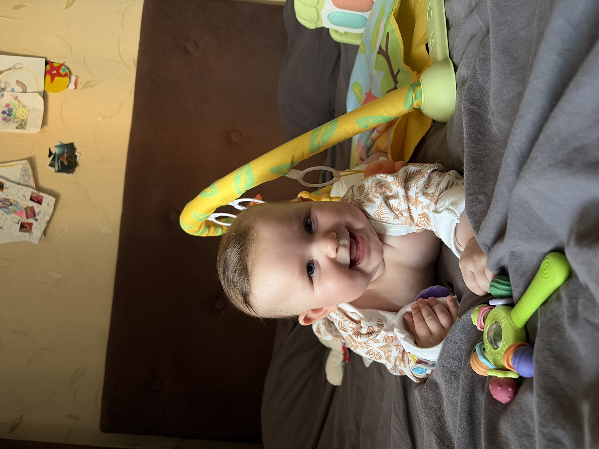 An infant is shown smiling brightly while lying on their stomach (tummy time) on a soft grey blanket. The baby, with light brown hair and blue eyes, is wearing a white long-sleeved top adorned with a light brown leaf pattern. Their head is propped up, looking directly at the viewer with a wide, happy grin.
Above the baby, a yellow and green play gym with a leafy fabric pattern arches over them, suggesting an indoor play area. To the baby's right (viewer's left), a colorful green rattle toy with various textured parts in orange, purple, and red rests on the blanket. In the baby's left hand (viewer's right), a soft purple toy is being held.
The baby is situated on a grey blanket that covers a dark brown upholstered surface, likely a bed or sofa. In the background, to the left, a light beige wallpaper with a subtle, swirling pattern is visible. Several children's drawings, a yellow star-shaped decoration, and small printed photographs are taped to this wall. The scene appears to be well-lit, indicating daytime. The overall impression is one of a happy baby engaged in playtime indoors.