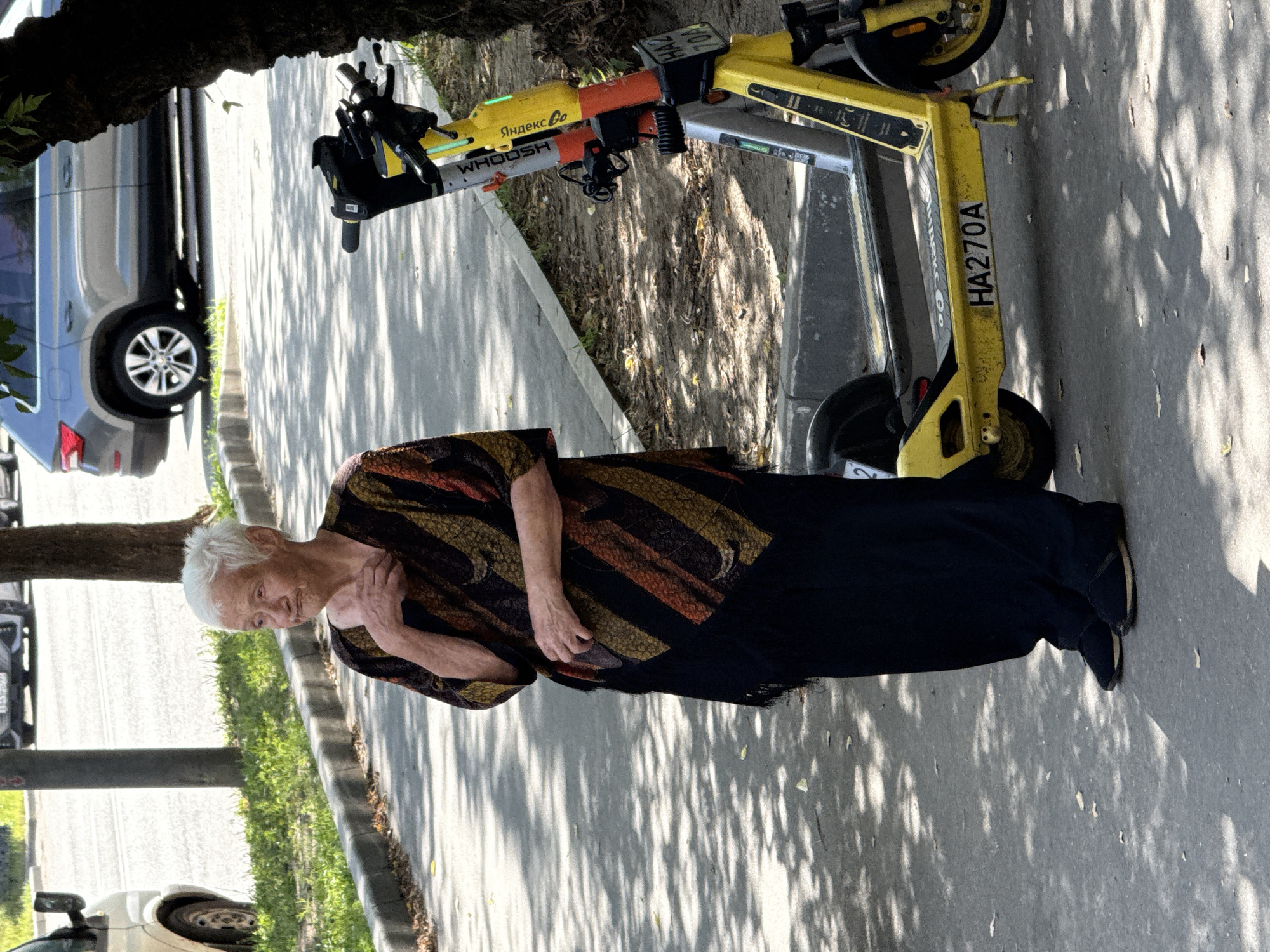 In Moscow, Russia, an elderly woman with white hair stands on a paved sidewalk, her right hand touching her neck. She is wearing a loose-fitting, patterned poncho with diagonal stripes of black, orange, and gold over dark pants. To her right, two yellow electric scooters are parked side-by-side. The scooter in the foreground has "HA270A" visible on its deck. A car is visible in the background on a street, and trees cast dappled shadows on the ground.
