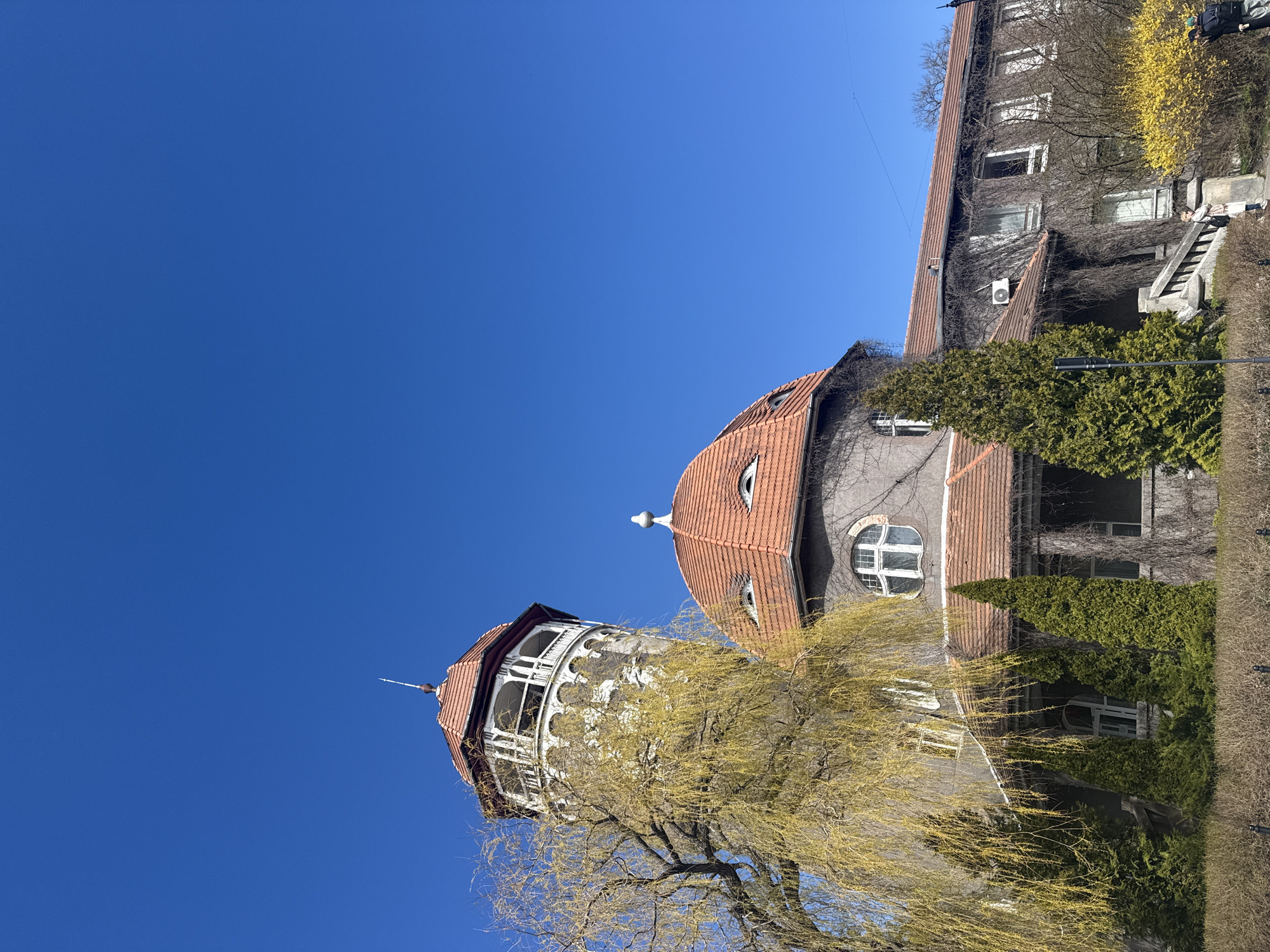 The image shows a water tower in Svetlogorsk, Russia. The tower is white and cylindrical with a red tile roof. Next to it is a building with a red tile roof and round windows. The buildings are surrounded by trees and bushes. The sky is clear and blue.