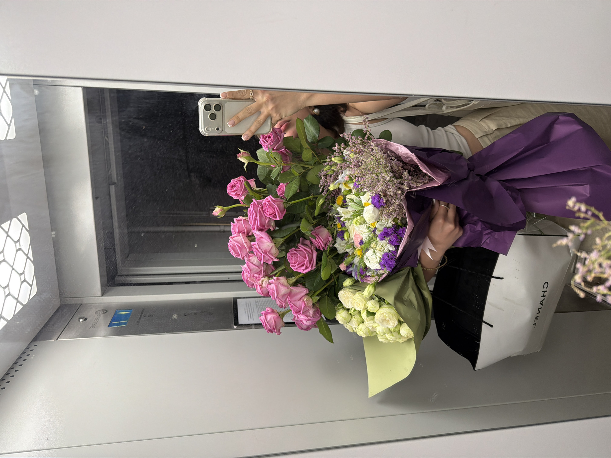 A mirrored selfie shows a person holding flowers and a shopping bag inside an elevator. The flowers are a mix of pink roses and white and purple blooms wrapped in purple and green paper. The shopping bag is white with black handles and the word "CHANEL" is written on it in reverse. The person is holding a smartphone to take the photo. The elevator's interior is light gray, and there's a window visible in the background. The location may be Yerevan, Armenia.
