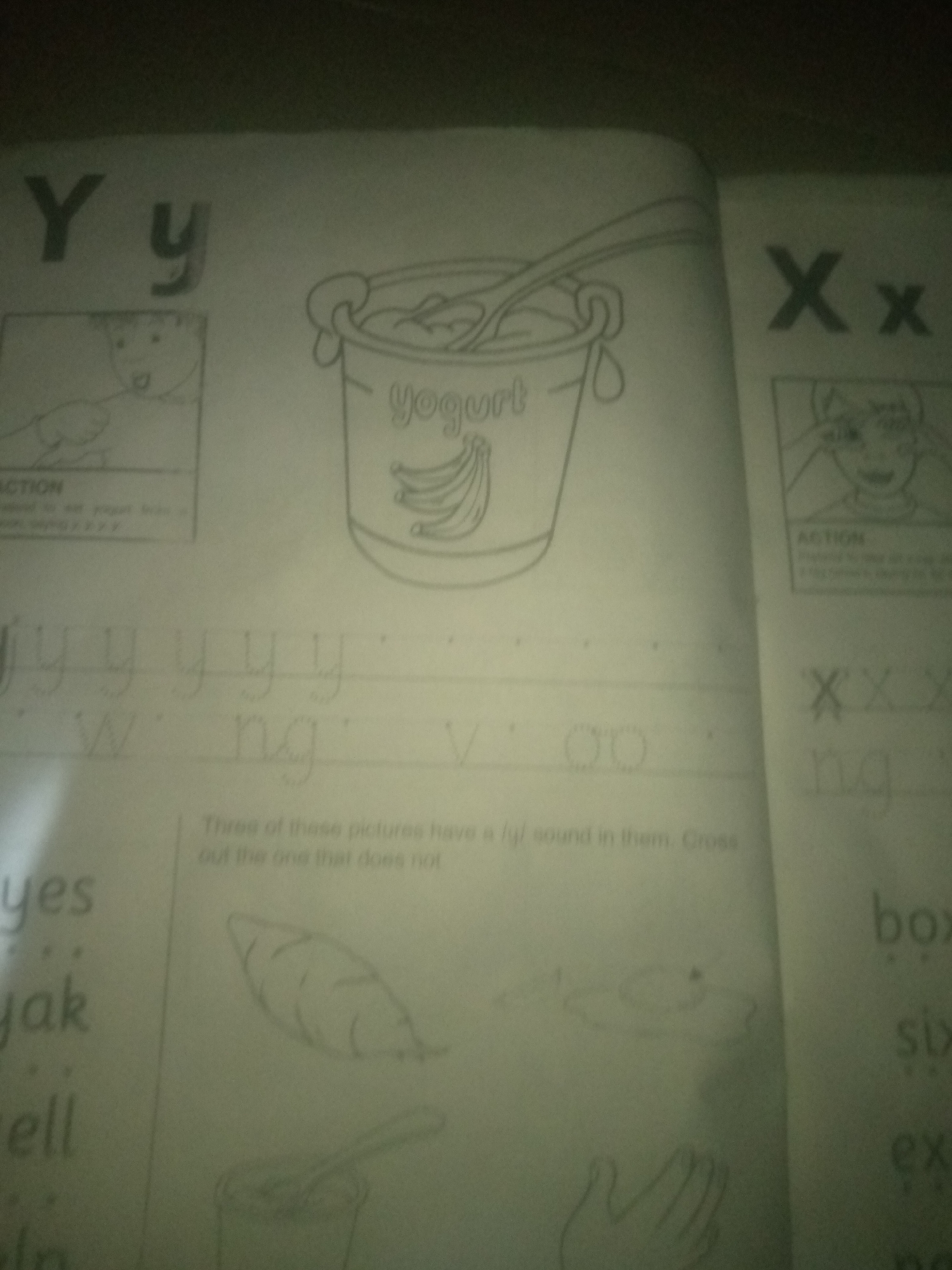 The image is a close-up of a workbook page, likely for young children learning the alphabet and vocabulary. The page is divided into sections with large letters and accompanying illustrations. On the left side, the letters "Y" and "y" are prominently displayed, followed by a drawing of a child eating with a spoon, labeled "ACTION." Below this, there are lines for practicing handwriting.
In the center of the page, a large drawing of a bucket of yogurt with a spoon in it is depicted, with the word "yogurt" written on the bucket. Below the yogurt illustration, there are more handwriting practice lines with letters like "w," "ng," and "v." Further down, a text box contains instructions: "Three of these pictures have a /y/ sound in them. Cross out the one that does not." Beneath this text are three small drawings: a carrot or yam, a fried egg, and a hand.
On the right side of the page, the letters "X" and "x" are shown, followed by a drawing of a person pointing to their ear, also labeled "ACTION." Below this are lines for handwriting practice, including the letters "x," "ng," and "box," "six," "ex," and "nx." The overall setting appears to be indoors, with the workbook laid out on a flat surface. The image is slightly blurry and the lighting is dim, suggesting it was taken in low-light conditions. There is no identifiable information about the location within Jauro Yinu, Nigeria.