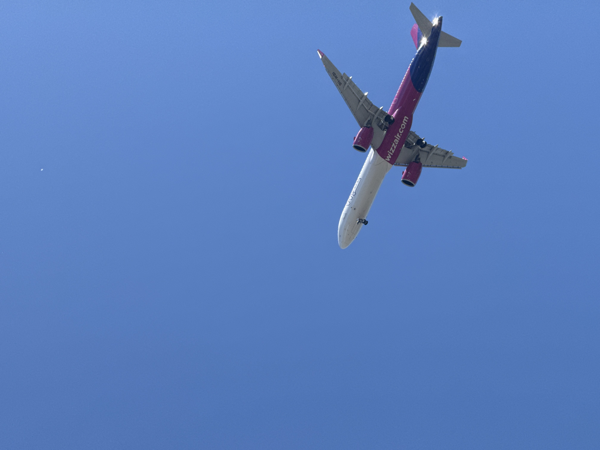 An airplane, identified as a Wizz Air Airbus A320 with registration HA-LXC, is captured from below against a clear blue sky. The aircraft is ascending or banking, with its underside predominantly visible. The distinctive magenta and blue livery of Wizz Air is prominent on the fuselage and tail. The text "wizzair.com" is clearly legible on the side of the aircraft. The engines, featuring a dark grey casing, are also visible. The lighting suggests it is daytime, likely mid-morning or afternoon, with no clouds present, indicating fair weather. The background is a uniform expanse of blue sky, offering no specific location cues other than the open atmosphere. There are no people visible, and the focus is entirely on the aircraft and the sky.