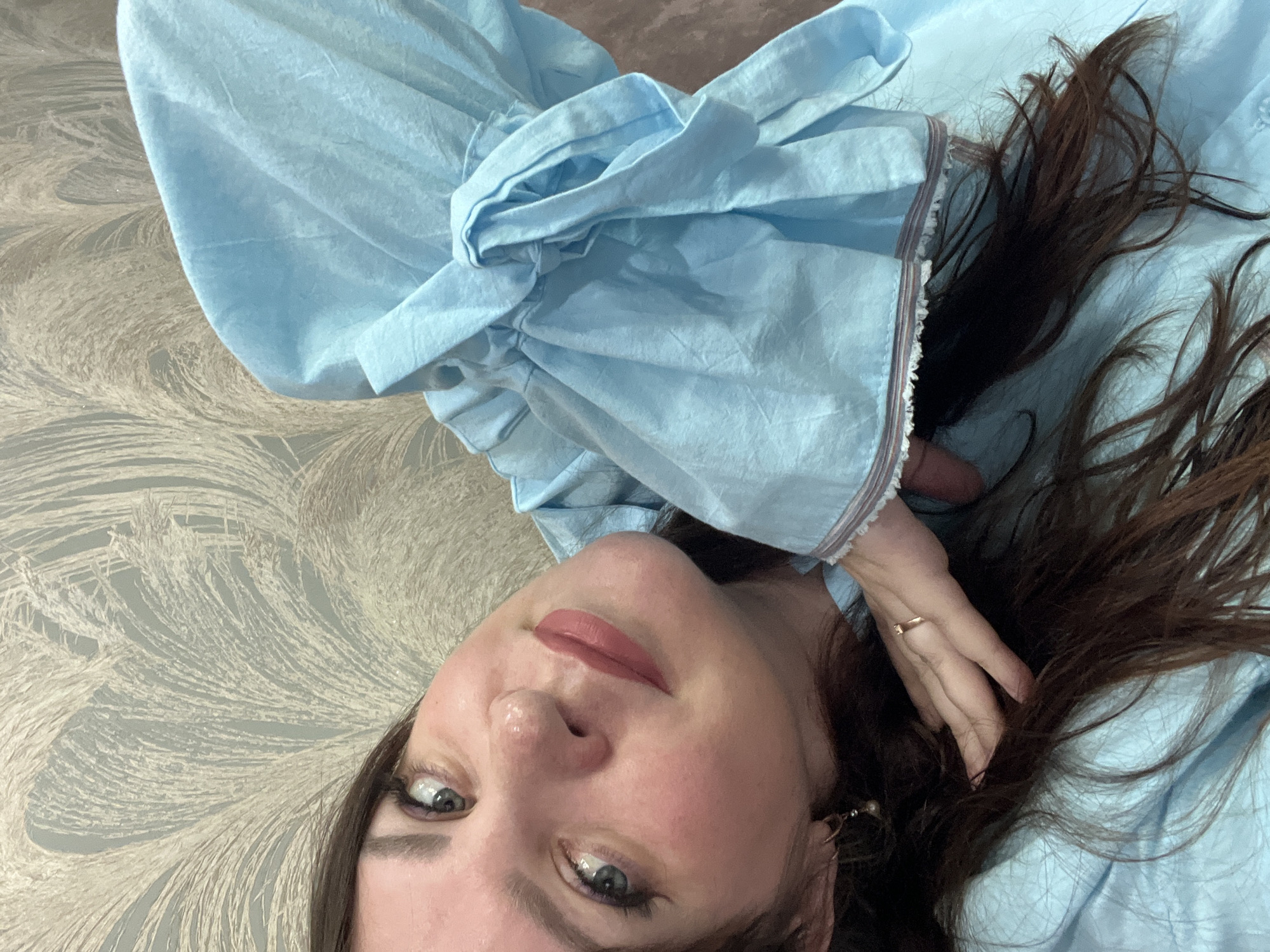 A selfie-style image features a woman with fair skin, brown hair, and blue-gray eyes looking up at the camera. She is wearing a light blue, ruffled blouse. Her hair is tousled and partially covers her right hand, which has a gold ring on one finger. Her makeup includes lipstick. The background consists of a patterned surface in beige and light gray tones. There is no indication of the city of Noviy, Russia, or any identifiable location.