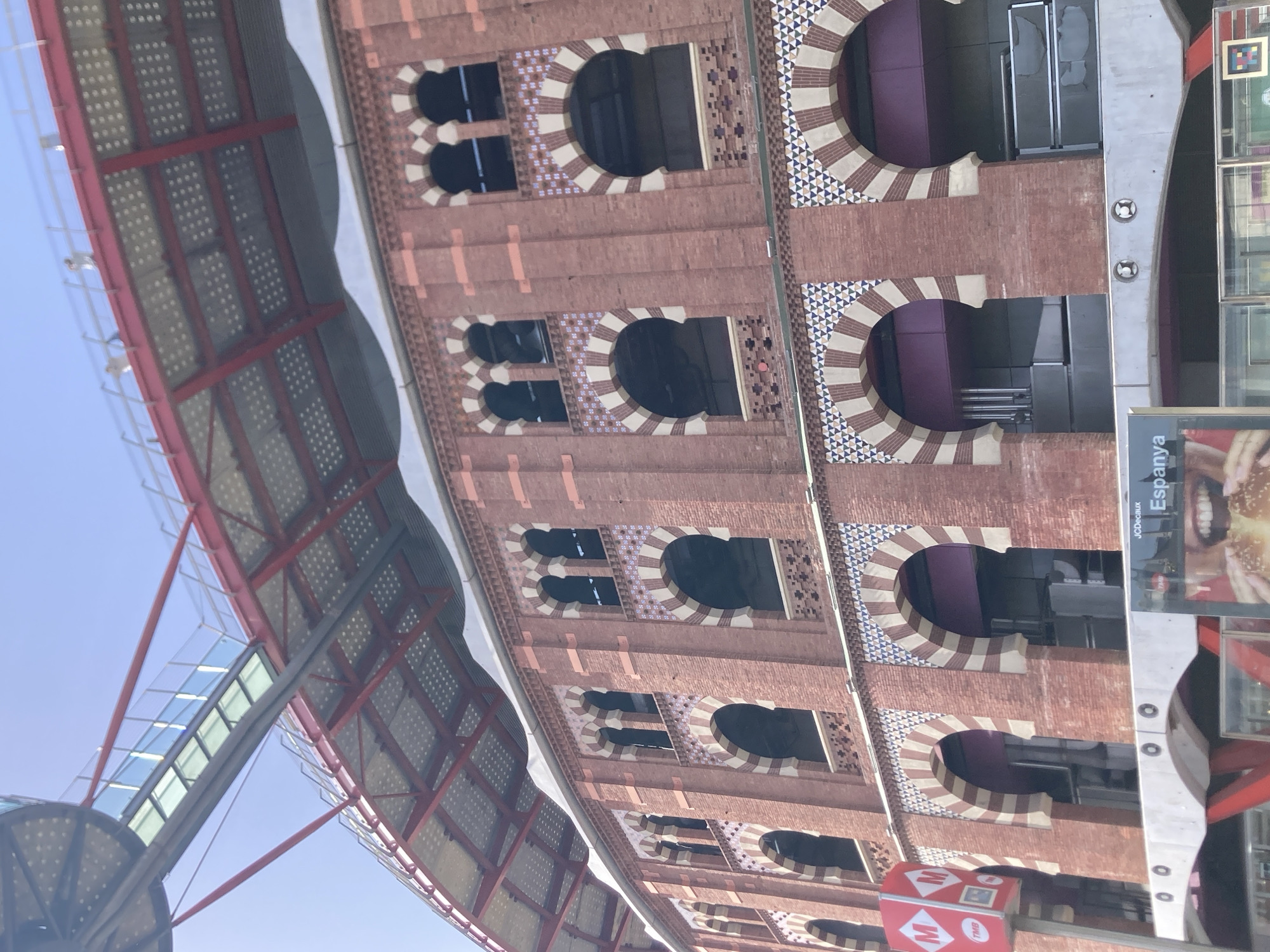 The image depicts the facade of the Las Arenas de Barcelona, a former bullring transformed into a shopping center, located in the Plaça d'Espanya, Barcelona, Spain. The building exhibits distinctive red brickwork with arched windows adorned with intricate geometric tile patterns in white, blue, and red. Above the main facade, a contemporary elevated walkway with a glass floor and red metal supports is visible, extending over the building. The sky above is a clear, light blue. In the foreground, a red sign with the white letter 'M' inside circles, likely indicating a metro station, stands to the left. A billboard displaying text and a close-up of a burger is partially visible at the bottom center. The overall scene captures the blend of historical architecture with modern urban infrastructure.