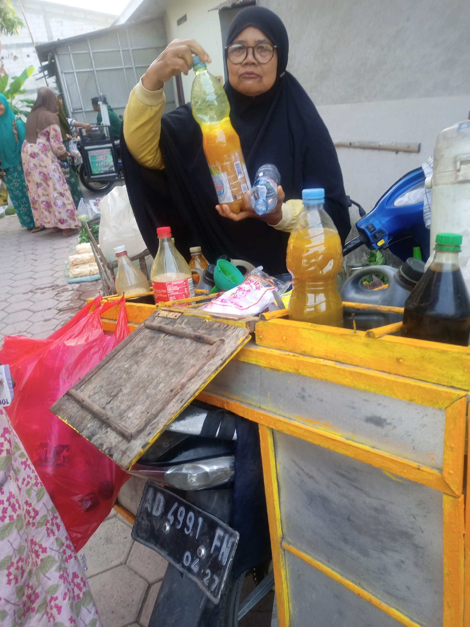 A middle-aged Indonesian woman wearing a black hijab and glasses stands behind a yellow food cart in the city of Nganti, Indonesia. She is holding up two plastic bottles, one with orange liquid and a label partially visible, and another smaller clear bottle. The cart is filled with various bottles of liquids, some red, some yellow, and some dark brown, as well as a red plastic bag and a wooden board. In the background, other people are visible, as well as a blue and black motorcycle. The ground is paved with hexagonal tiles. A license plate with the text "AD 4991 FN" and "04.27" is attached to the cart.