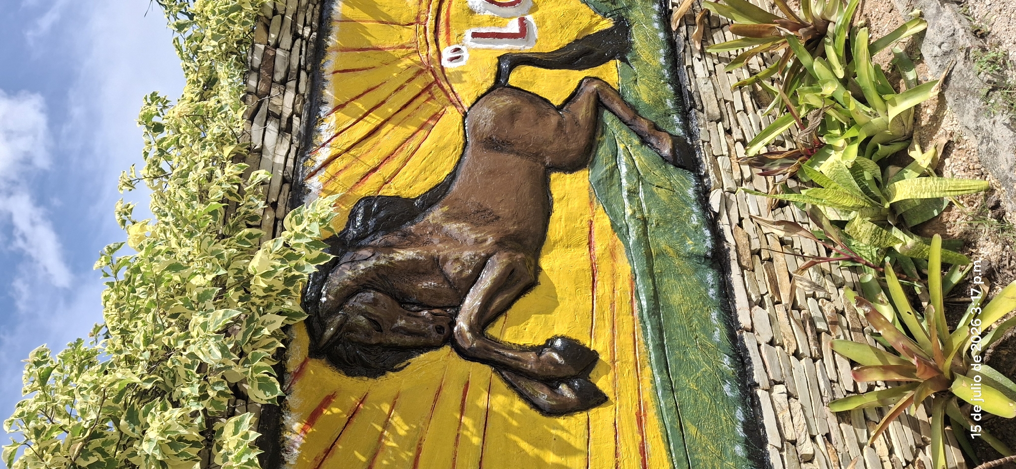 In the city of Vallecito, Venezuela, a vibrant mural adorns a stone wall. The mural depicts a brown horse rearing up against a bright yellow sunburst. The horse is rendered in a bold, three-dimensional style, with its body textured to mimic muscle and mane. Below the horse, a textured green landscape adds depth to the scene. The mural is partially obscured by lush green and yellow foliage cascading from above, and a stone wall forms the base of the artwork. In the foreground, various tropical plants with pointed leaves are planted in the sandy soil. A timestamp in the lower-left corner reads "15 de julio de 2026 3:17 p.m."