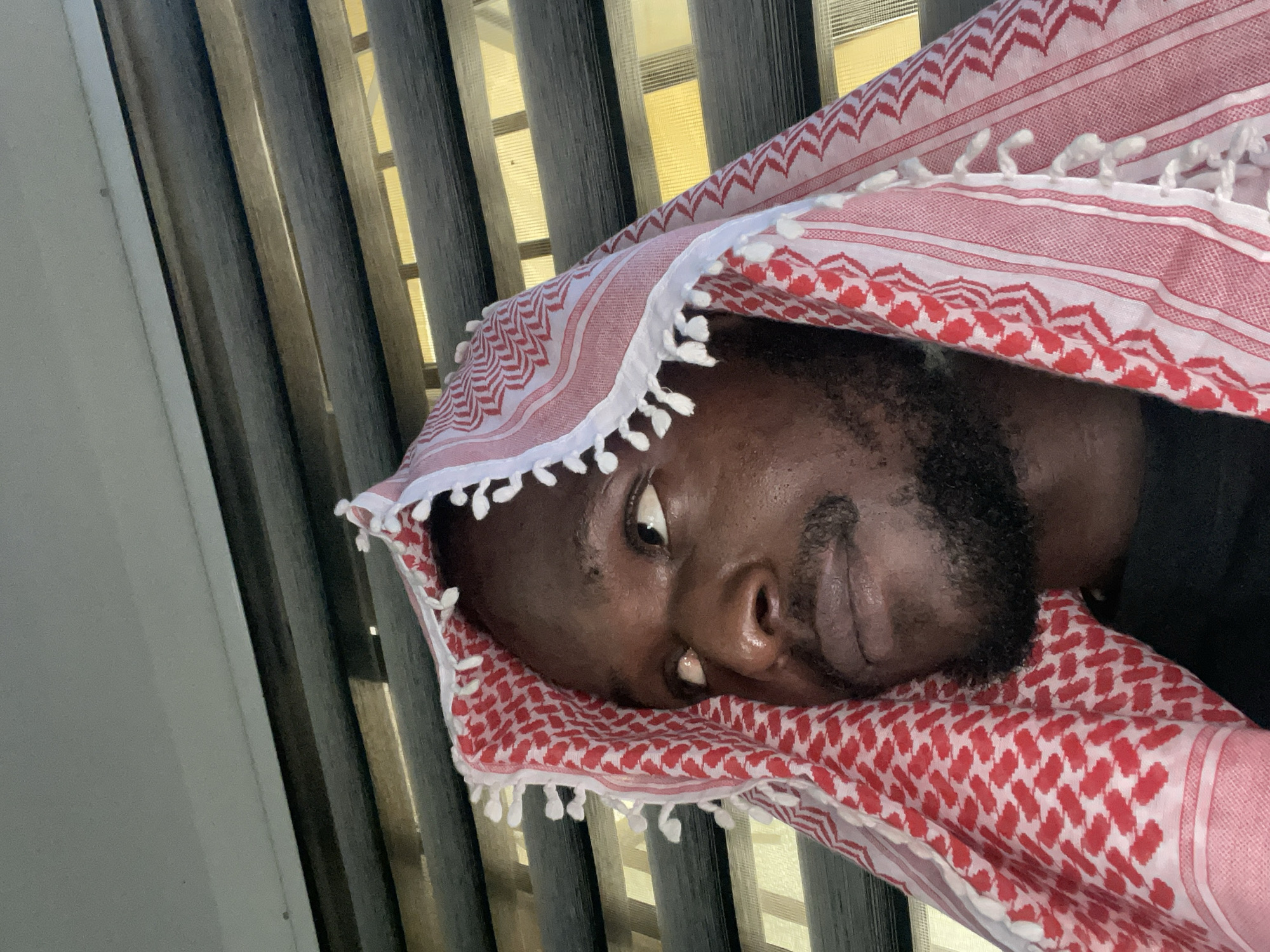 A medium shot, from a low angle, shows a man wearing a red and white keffiyeh. He is looking up and to his left, with his face partially obscured by the keffiyeh and the light from behind. The keffiyeh has a white fringe along its edges. Behind him are horizontal blinds with light visible through the slats. The image is likely taken indoors, possibly in a room with windows covered by blinds. The man's facial features, including his eyes, nose, and mouth, are visible. He has a beard and mustache. The background suggests a domestic or office environment, common in cities like Abuja, Nigeria.