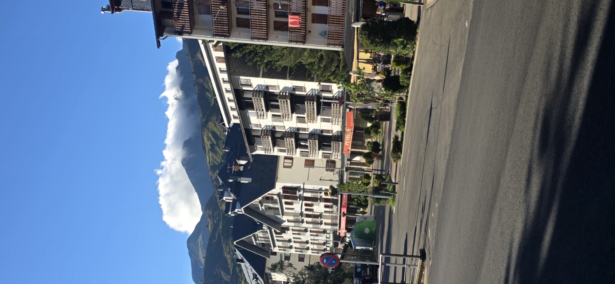 This is a vertical, eye-level, outdoor, daytime, medium-long shot of a street in Sallent de Gállego, Spain, with a backdrop of mountains. The scene features several multi-story buildings lining the street. The buildings are modern in style, with balconies and dark pitched roofs. Some of the buildings have businesses on the ground floor, indicated by awnings and outdoor seating. One awning prominently displays the word "PETRUSO". In the foreground, there is an asphalt road with shadows cast across it. To the left of the road, a metal barrier and a circular sign with a red border and a blue and red diagonal line are visible. Further down the street, a green recycling bin and parked vehicles can be observed. The background consists of rolling, green-covered mountains partially obscured by a fluffy white cloud formation. The sky is a clear, bright blue. There are no people clearly visible in the image, though some figures are suggested at outdoor seating areas of the businesses.