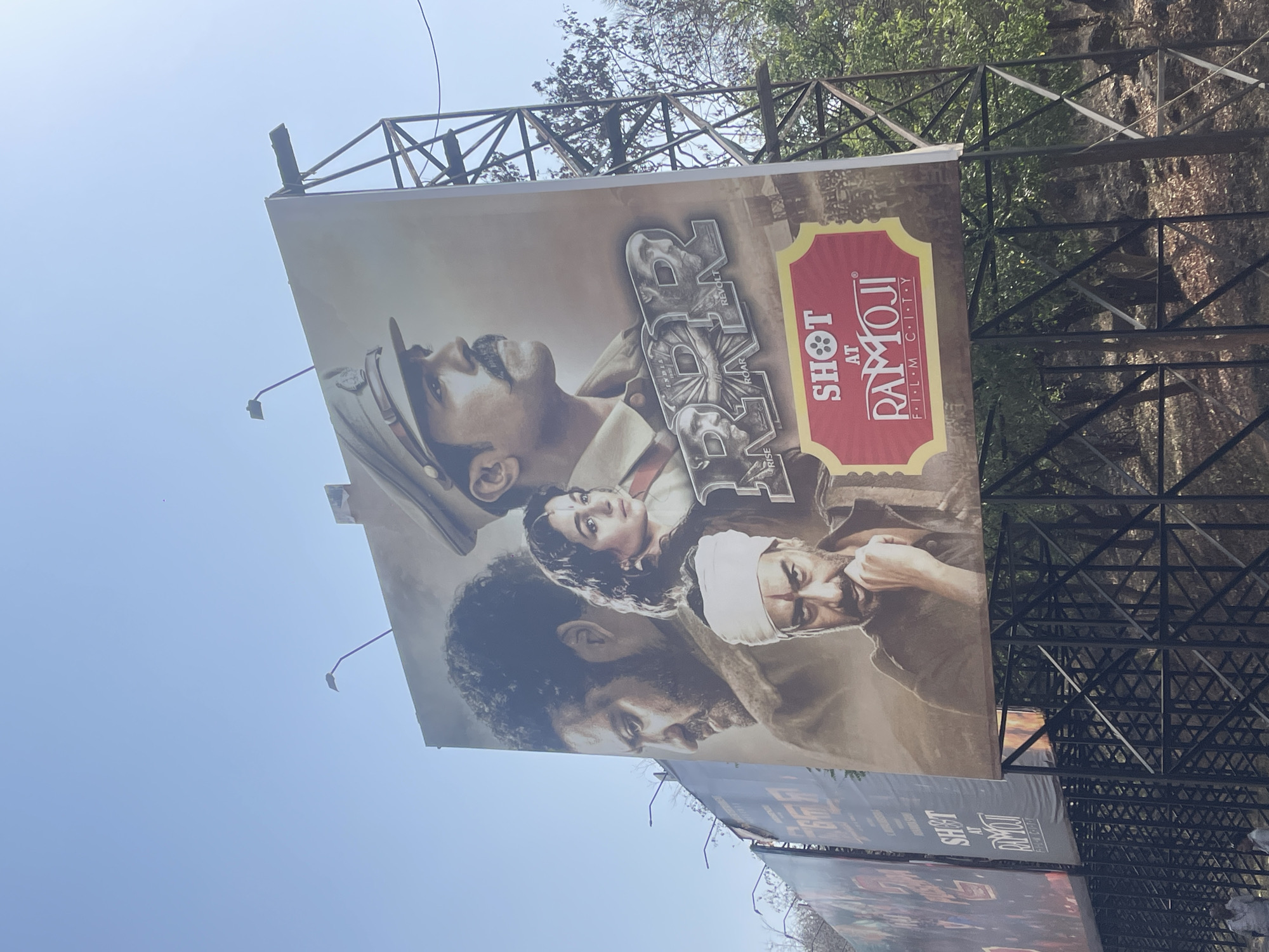 A low-angle, outdoor photograph shows a billboard advertising the film "RRR," shot at Ramoji Film City, located somewhere in India. The billboard is mounted on a metal frame structure. The sky is blue. The billboard features a collage of the movie's actors. Below the "RRR" billboard, another billboard is visible. Trees and foliage can be seen behind the billboard and its support structure.