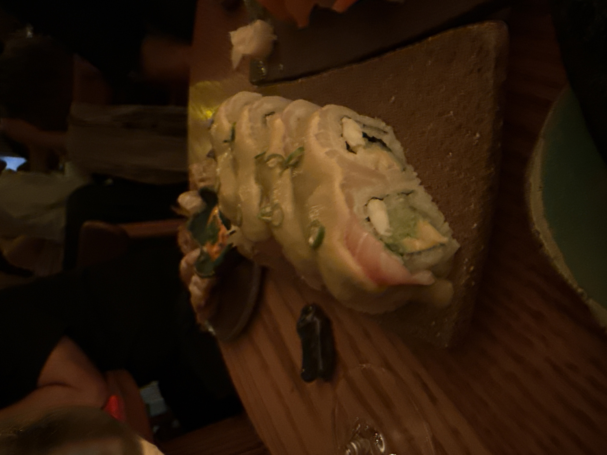 A close-up shot depicts a plate of sushi on a wooden table. The sushi is rolled and covered in a light sauce, with some green garnish sprinkled on top. A small amount of ginger is visible on the plate. A small piece of black object, possibly a utensil or condiment, is also on the table near the sushi. The lighting is dim, creating a warm, intimate ambiance. Other people are present in the background, with one visible arm. The photograph could be taken in Mexico City, Mexico.