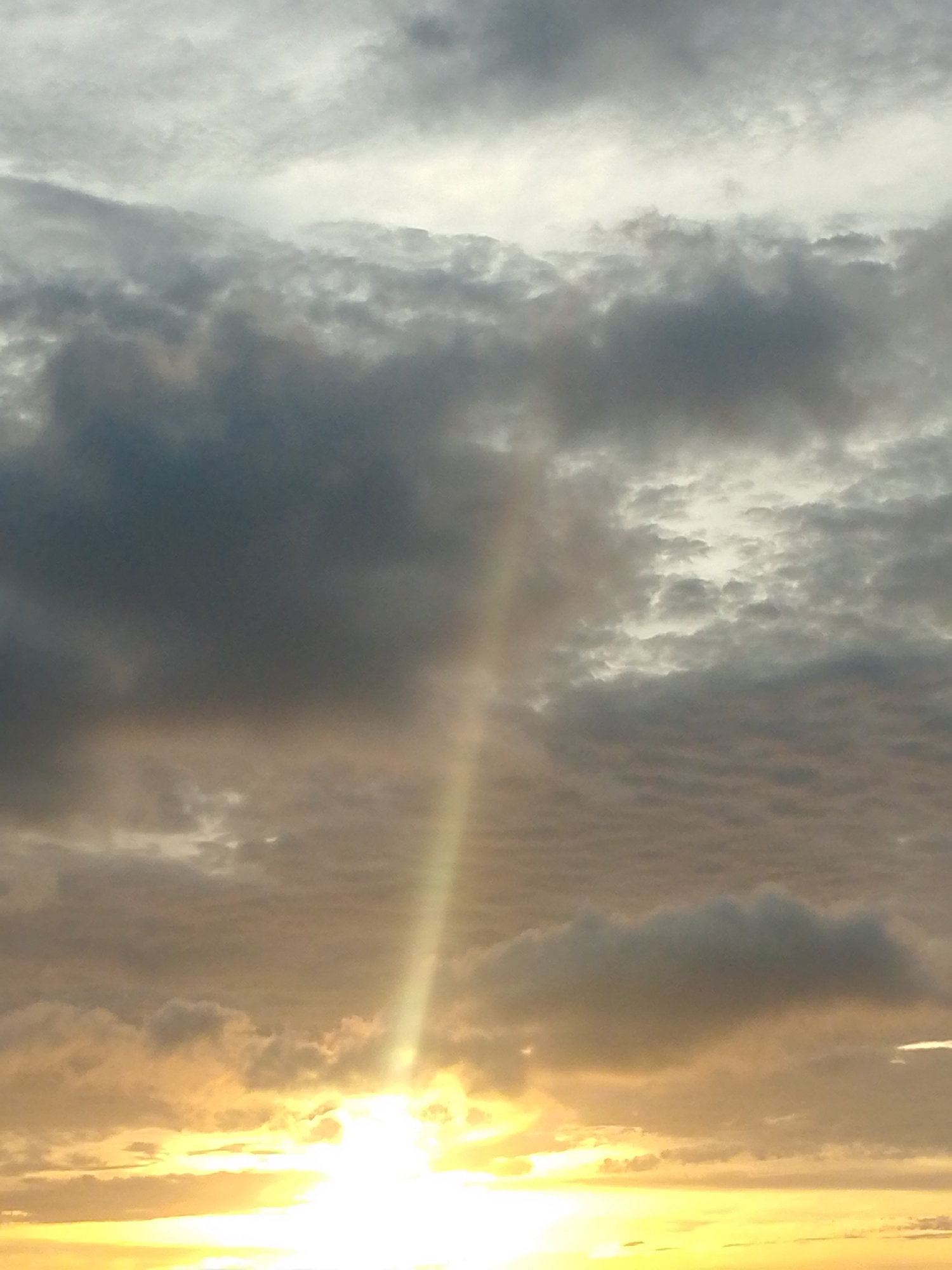 The image shows a bright, golden sunset in Juan Griego, Venezuela. A single, prominent ray of sunlight shines vertically through a break in the cloud cover. The sky is filled with layers of clouds, ranging from dark gray to lighter white hues. The sun itself is visible near the horizon, obscured by some cloud cover but creating a bright, radiant glow. The horizon appears to be over water.