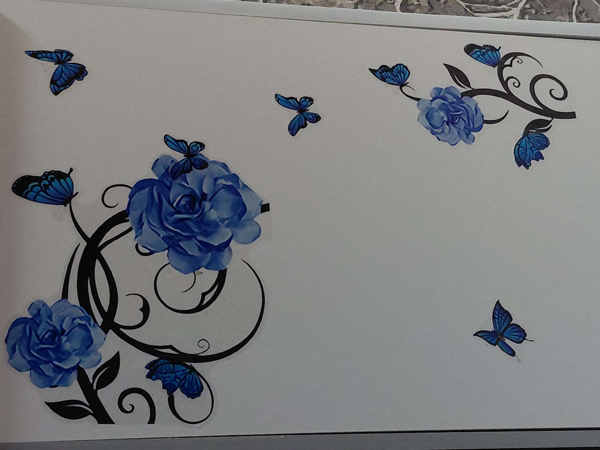 The image displays a decorative arrangement of blue butterflies and blue roses with intricate black swirling patterns, set against a plain white background. Three primary blue roses are visible, varying in size and placement. The largest rose is located in the bottom left corner, accompanied by extensive black swirling vines and a blue butterfly with black outlines. Another prominent blue rose is positioned in the upper middle-left, with a smaller blue butterfly appearing to rest on its petals. A third blue rose, also connected to black swirling elements, is featured in the upper right, surrounded by two blue butterflies.
Several individual blue butterflies are scattered across the white surface: one in the top left, one in the top middle, and one in the bottom right. The butterflies exhibit varying shades of blue, from a vibrant cerulean to deeper indigo tones, with black details outlining their wings. The roses are rendered in a soft, watercolor-like style, showcasing different shades of blue for depth. The black swirling patterns provide a contrasting, elegant framework for the floral and insect elements.
The overall design appears to be a decal or a print on a flat surface, such as a wall or a decorative panel. The background is uniformly white, with a subtle hint of a textured edge along the very top, possibly indicating a wall or a framed object. No people, discernible activities, or visible text are present in the image.