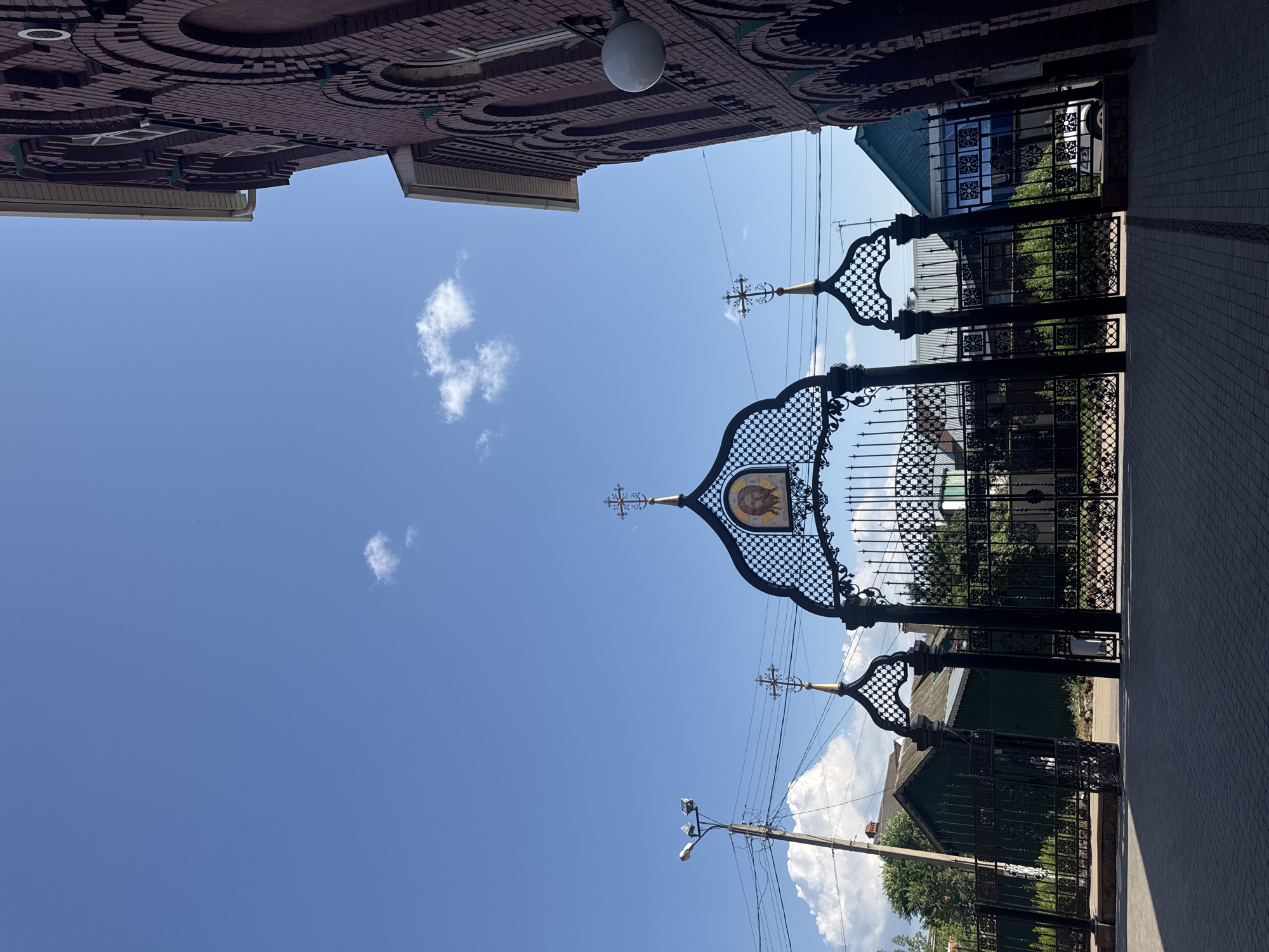 The image depicts an ornate black metal gate, featuring decorative arches and crosses, leading into a courtyard. Above the central arch, an icon of Jesus is visible. To the right, a portion of a red brick building with arched windows and decorative brickwork is shown. The sky is a clear blue with a few scattered white clouds. In the background, green trees and the roofs of houses can be seen. A lamppost with lights is positioned to the left of the gate. The ground in front of the gate is paved with grey tiles. This scene is located in the city of Tolyatti, Russia, specifically at the Church of the Kazan Icon of the Mother of God.