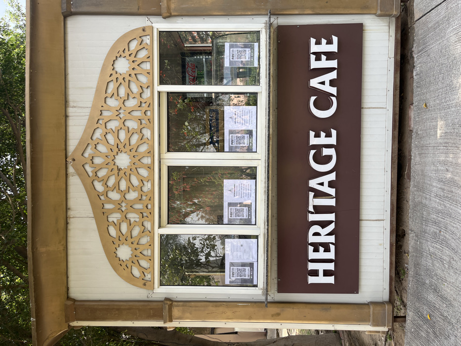 A small, white, standalone kiosk with a brown awning and a brown, rectangular sign that reads "HERITAGE CAFE" in large, white letters. The sign is directly above a window with three glass panes, each containing a variety of advertisements. The kiosk is situated in the city of Sahibzada Ajit Singh Nagar, India. The location is surrounded by green trees and a paved area.