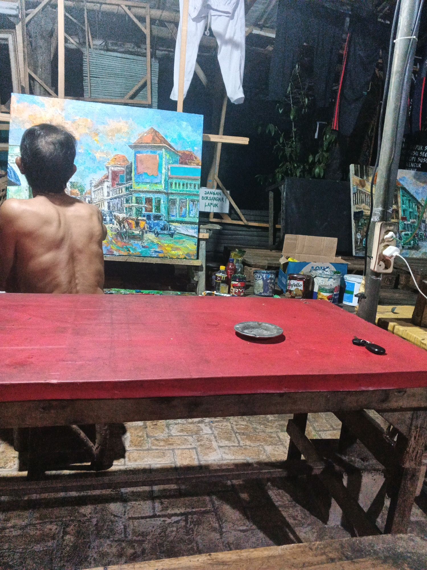 In Ungaran, Indonesia, an artist is seen from behind, shirtless and facing a large, vibrant oil painting. The painting depicts a street scene with colorful buildings, cars, and a horse-drawn carriage under a bright, cloudy sky. The artist is positioned in front of an easel, with the painting displayed prominently.
The setting appears to be an outdoor workspace, possibly a studio or a covered area. A bright red table occupies the foreground, with an ashtray and a dark object, possibly a pen or a tool, placed on its surface. Various items are scattered on the table behind the artist, including jars, bottles, and what appears to be a box of pasta or food. A white cloth hangs from a wooden frame in the background.
The ground is paved with what looks like light-colored bricks or tiles. The overall lighting suggests it might be late afternoon or evening, with shadows cast across the scene. The environment seems somewhat rustic, with wooden structures and what looks like corrugated metal roofing visible in the background, hinting at a humble or informal artistic space.