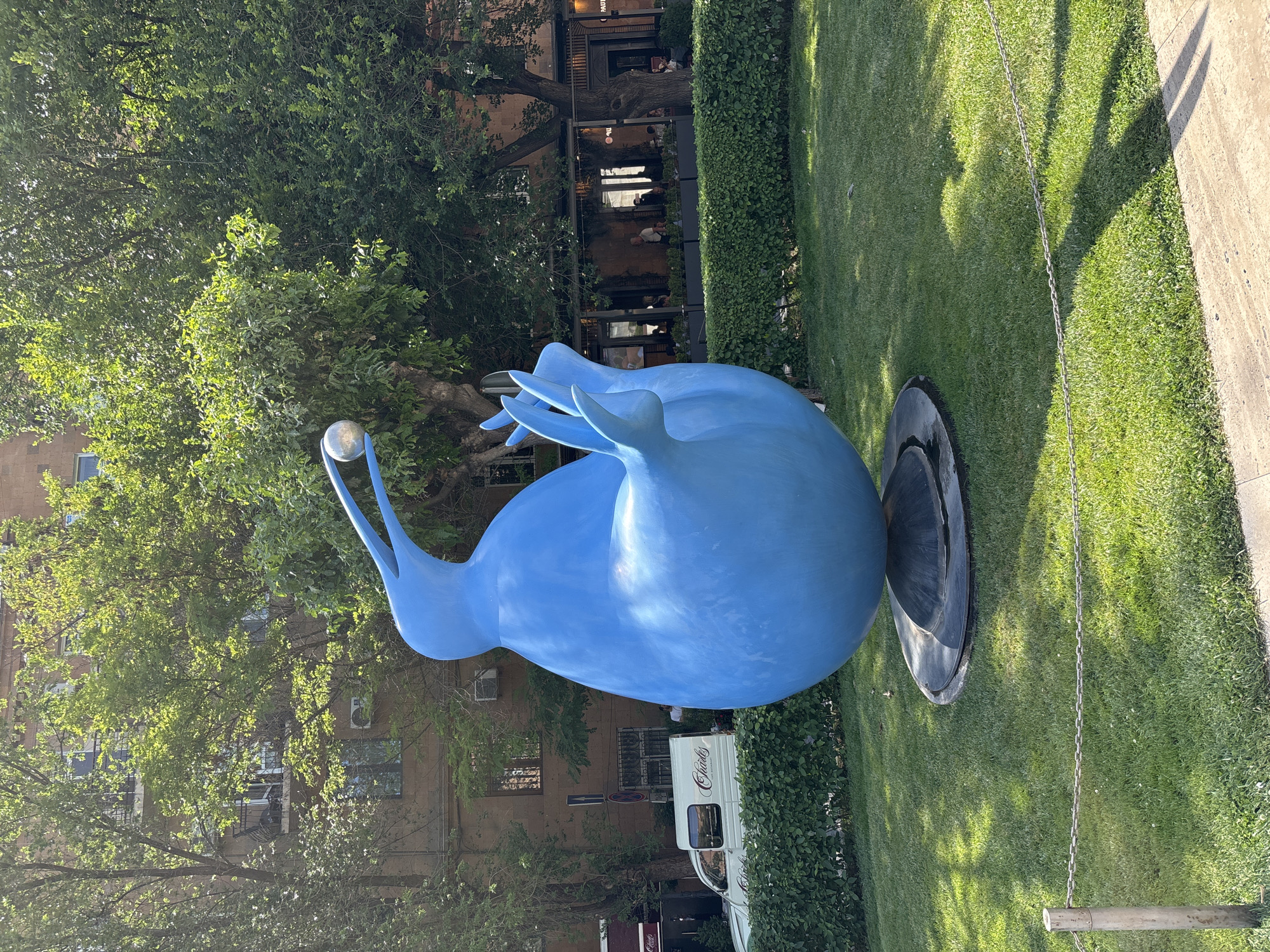 This image depicts a large, bright blue, abstract sculpture of a bird-like creature holding a silver sphere. The sculpture is situated on a circular, dark grey base within a well-maintained green grassy area, bordered by a chain fence and a paved walkway in the foreground. In the background, a building with brown brick facade, balconies, and trees with lush green foliage are visible. To the left of the sculpture, a white van with "Charles" written on its side is parked. Further back, a street with outdoor seating areas of restaurants and people can be observed. This scene is located in Yerevan, Armenia, specifically in front of the Saryan Street building.