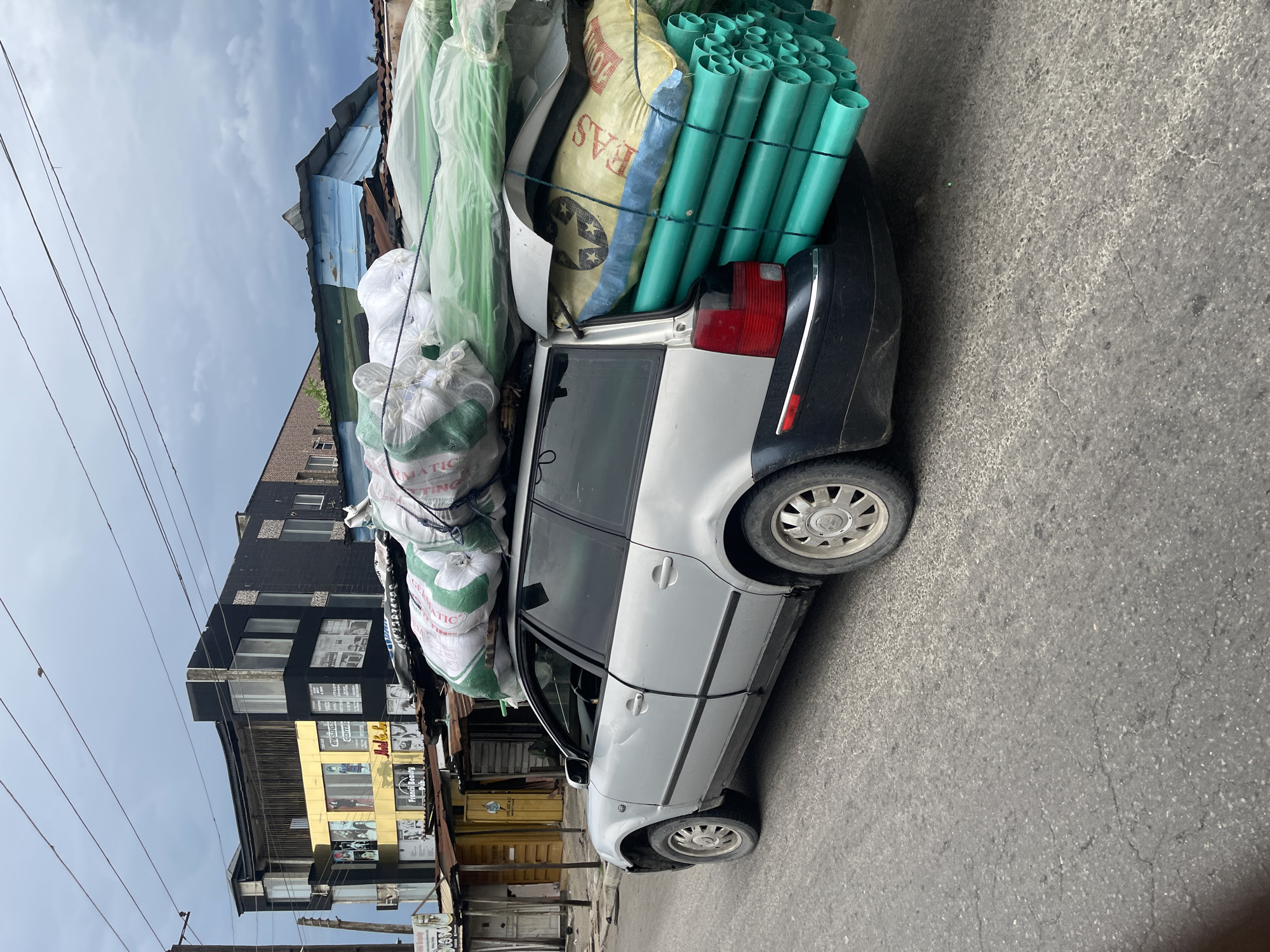 A silver station wagon, seemingly a Volkswagen Passat, is severely overloaded with goods. The vehicle is packed with green PVC pipes, large sacks, and other materials wrapped in plastic. The car's suspension is heavily compressed due to the weight.
The scene appears to be on a street in Ado, Nigeria. The background includes buildings, some with commercial signage. Power lines are visible in the sky. The road is paved but shows signs of wear and tear. There are no visible people actively interacting with the vehicle.