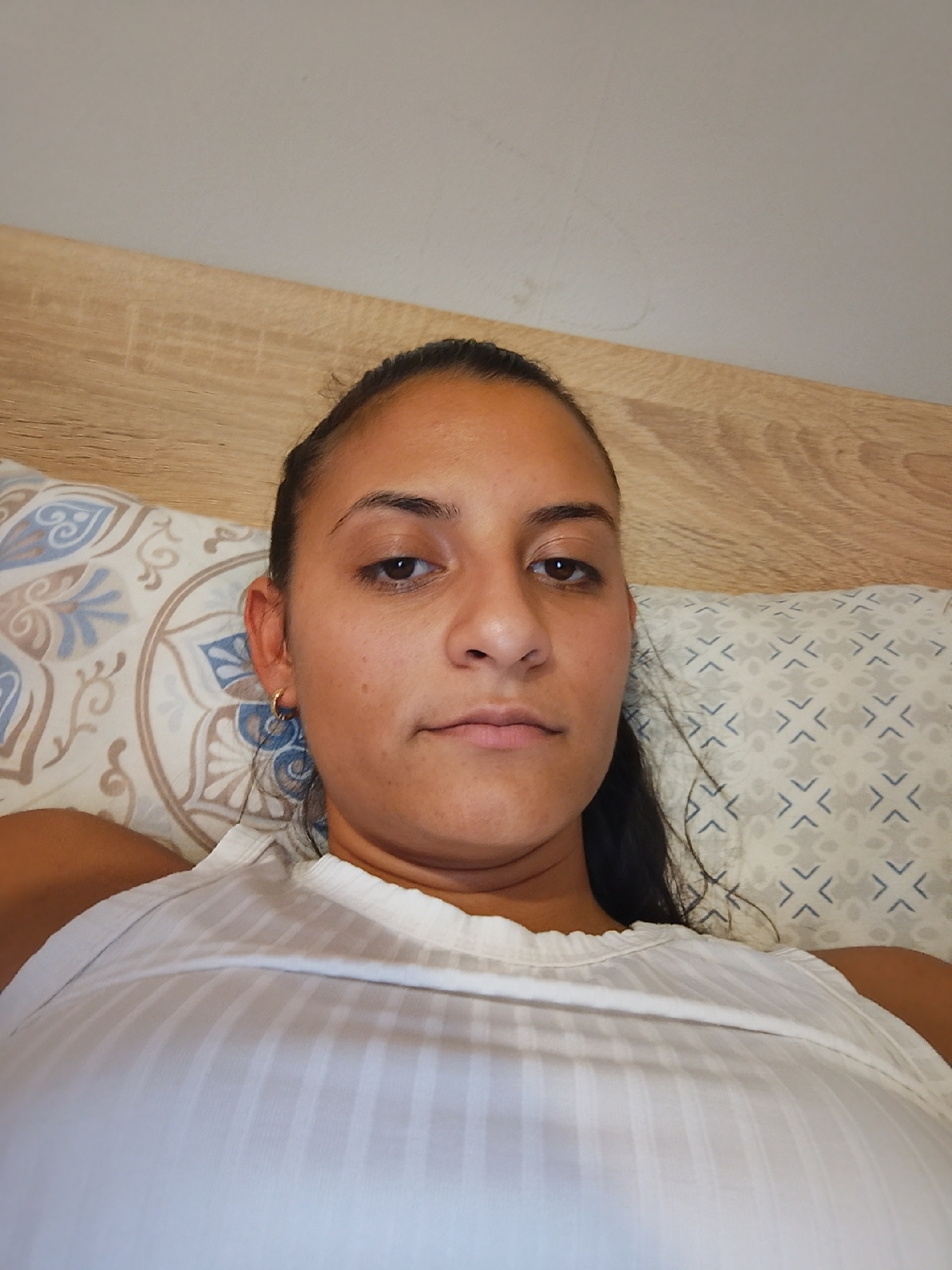 The image shows a close-up, over-the-shoulder selfie of a woman lying in a bed. She has dark hair pulled back and is wearing a white, ribbed tank top. Her face is the primary focus, with her eyes looking directly at the camera. She is wearing a small gold hoop earring in her visible ear. The background consists of a wooden headboard and pillows with patterned covers. The scene appears to be indoors, in a bedroom setting. There is no visible indication of the location being in the city of Yambol, Bulgaria.