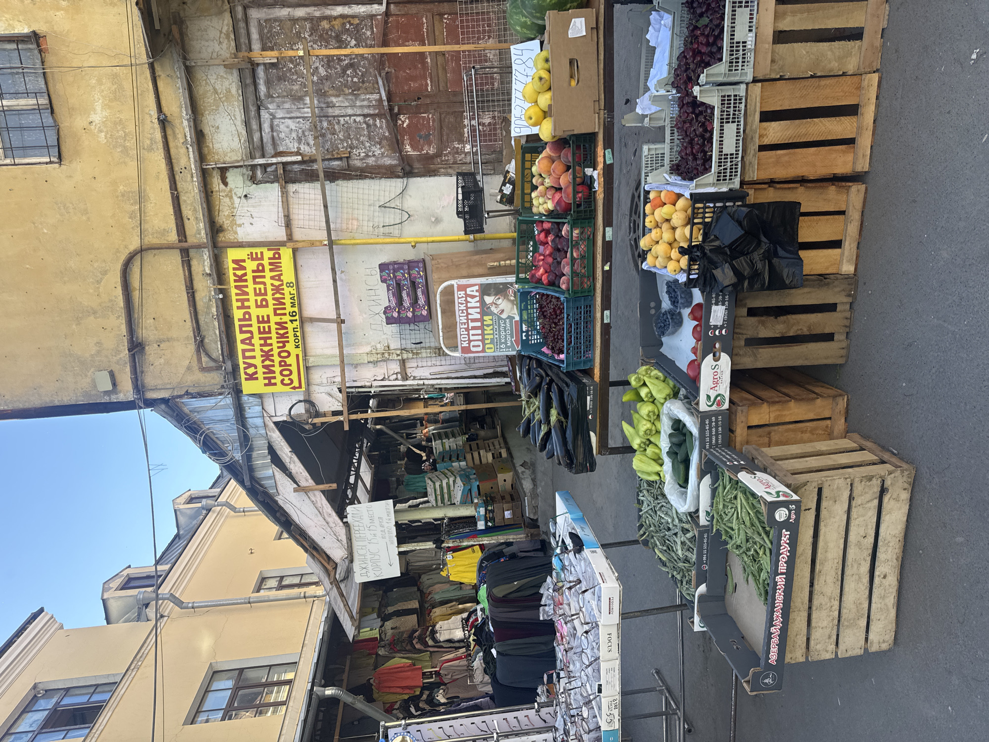 This image depicts an outdoor market scene, likely in Saint Petersburg, Russia, given the Cyrillic text. The camera is positioned at an angle, making it appear as though the scene is tilted. The foreground features stalls selling fresh produce, including eggplants, peppers, green beans, blueberries, grapes, apricots, and apples, displayed in wooden crates and plastic containers. There are also stalls selling clothing, including swimwear, underwear, shirts, and pajamas, as well as a stall with eyeglasses.
Several signs are visible, one in yellow advertises "купальники нижнее бельё сорочки пижамы" (swimwear, underwear, shirts, pajamas) and gives a location "корп. 16 маг. 8" (corp. 16 shop 8). Another sign, in white with red lettering, advertises "корейская оптика" (Korean optics) and "очки" (eyeglasses). A white sign with Cyrillic text states "ДЖИНСЫ ПЕРЕЕХАЛИ КОРПУС 15 15 МЕСТО" (Jeans moved to building 15, 15th spot). Some of the produce crates also have text, including "АЗЕРБАЙДЖАНСКИЙ ПРОДУКТ" (Azerbaijani product) and the brand "Agro S".
The background shows the facade of an older building with peeling paint and weathered wooden doors. The sky is visible, suggesting it is daytime and likely clear weather. There are no people clearly discernible in the image, though the presence of market stalls implies a commercial setting where people would be shopping and selling. The overall impression is a bustling, slightly chaotic market environment.