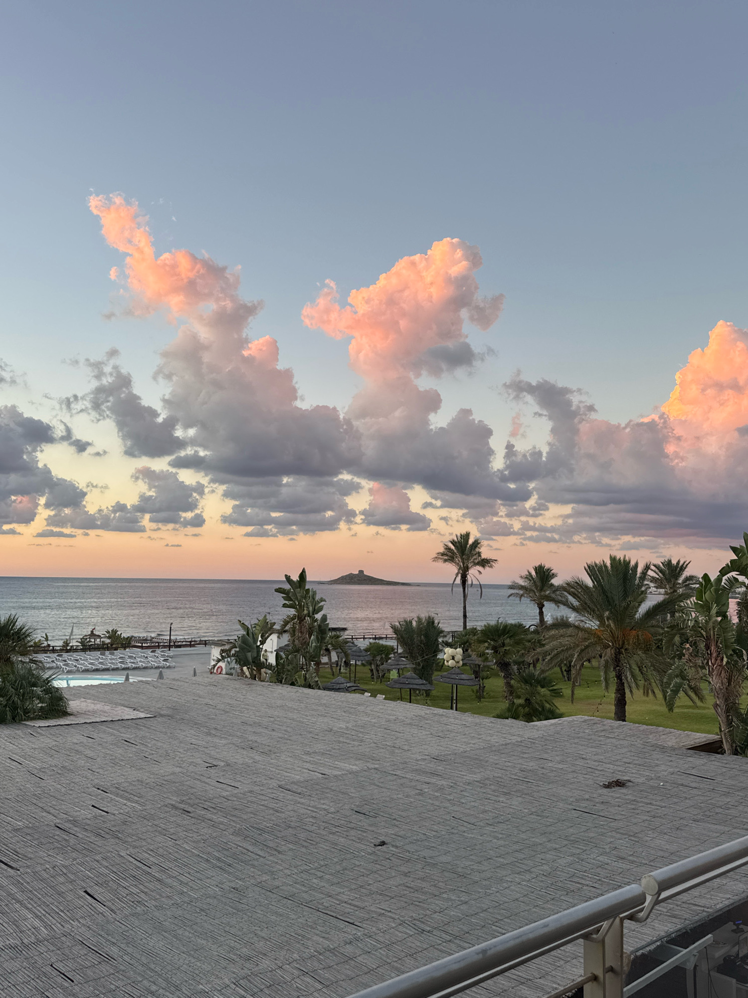 This is a landscape photograph taken at dusk from a raised vantage point, likely a balcony or rooftop, overlooking a coastal resort area. The foreground is dominated by a textured surface, appearing to be a woven mat or thatched roof, with a metal railing in the lower right corner. Beyond this, a swimming pool with loungers is visible, followed by a stretch of beach with more loungers and umbrellas. The sea extends to the horizon, where a small island with a structure on top is discernible. The sky is filled with dramatic clouds, illuminated by the setting sun, casting shades of pink, orange, and grey. Palm trees and other lush greenery are scattered throughout the scene, framing the view of the sea and the island. The overall impression is one of a serene, tropical evening. The location is identified as Isola delle Femmine, Italy, suggesting this view is from a hotel or resort situated near the iconic island.