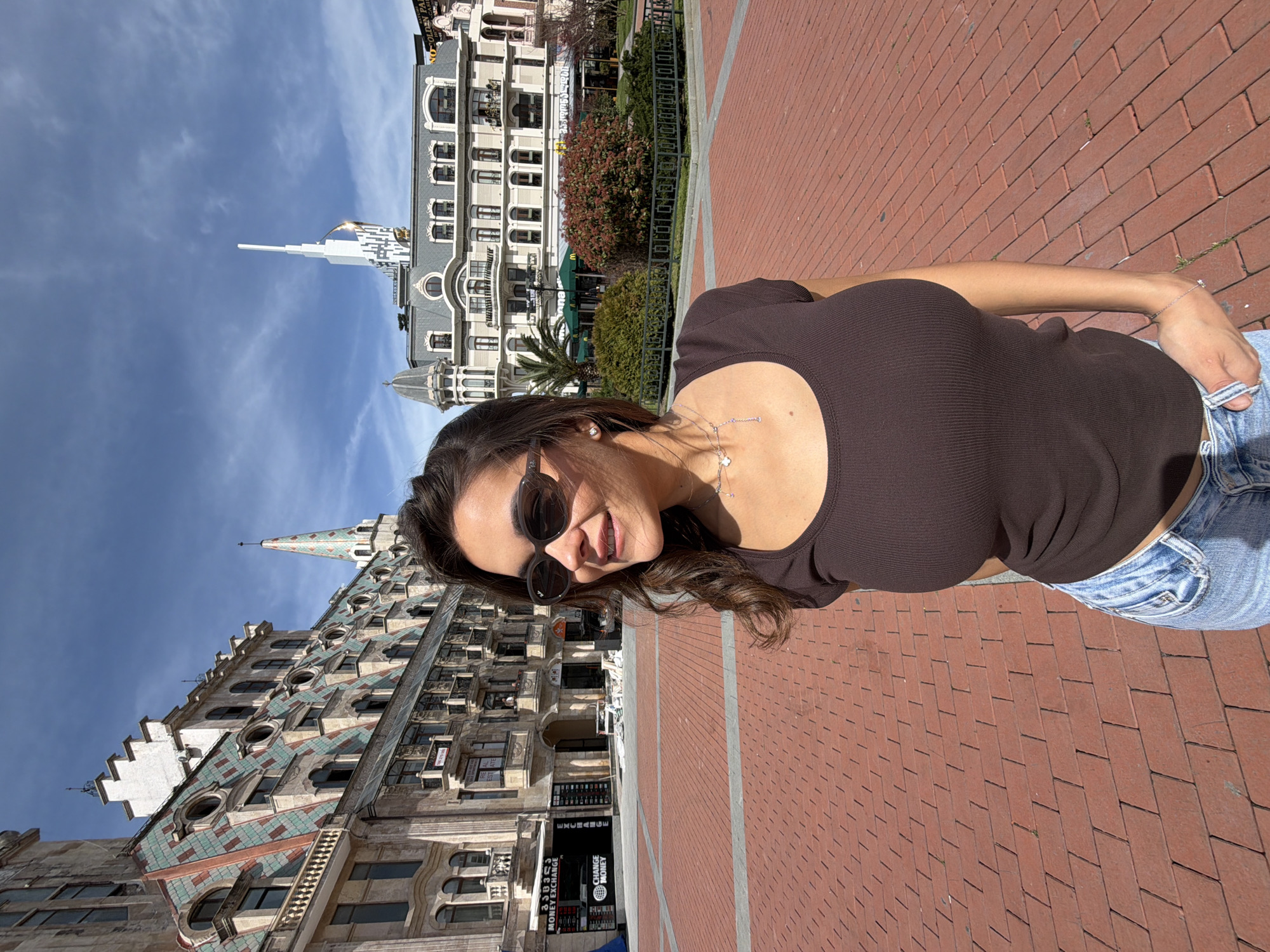 A woman in sunglasses and a brown shirt stands in a public square in Batumi, Georgia. In the background are two distinct buildings: one with a white spire and a modern design and the other a classical building with a brown roof. The ground is paved with red bricks.