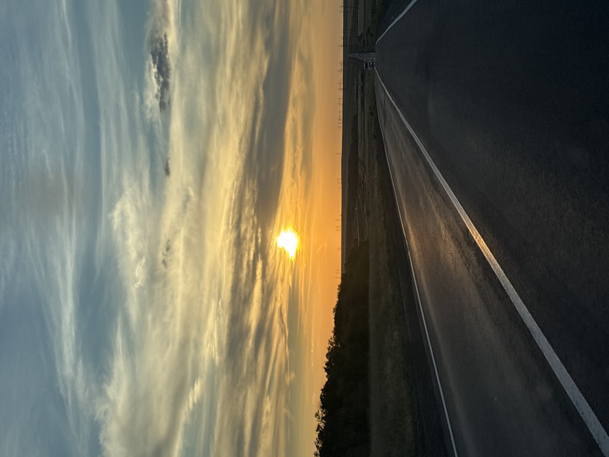 This is a vertical, full shot of a road stretching into the distance towards a sunset in Қазақ, Kazakhstan. The sky is filled with dramatic clouds in hues of blue, grey, and orange, with the bright sun appearing as a glowing orb on the right side of the frame. The road, made of asphalt, has white lines marking its edges and center, and it reflects the warm sunset light. On the left side of the road, there is a dark, dense line of trees. In the distance, the landscape appears flat, with rows of wind turbines visible against the horizon. A single truck is seen on the road, traveling away from the viewer, contributing to the sense of a journey. The overall atmosphere is serene and vast, capturing the essence of a rural landscape at dusk.