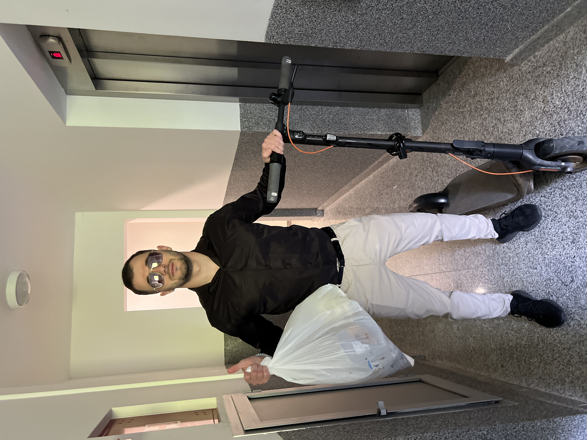 A man is standing in an interior hallway near an elevator. He is wearing sunglasses, a black jacket, white pants, and black shoes. He is holding a white plastic bag in his left hand and a black electric scooter in his right hand. The hallway has a gray tiled floor and off-white walls. There is a metal door and an elevator with a lit-up button indicating floor level. There is no indication that this scene is in Boreti, Montenegro.