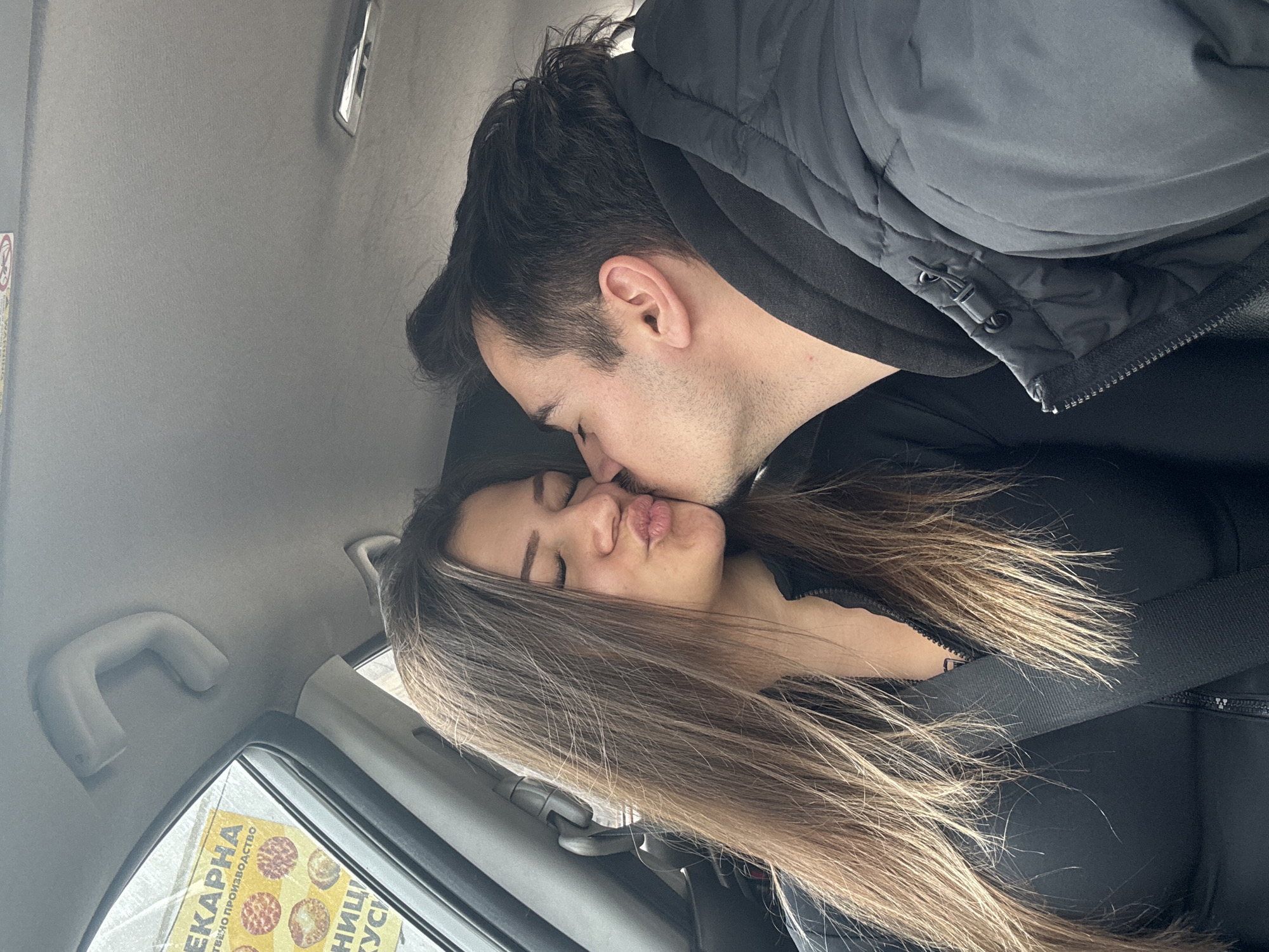 A photograph taken inside a vehicle depicts a man kissing a woman on the lips. The woman has long, light brown hair. Both are wearing black clothing. The scene suggests they are inside a car. A yellow poster with Bulgarian text and pictures of food can be seen through the car window. Given the text language, the location is likely Sofia, Bulgaria.
