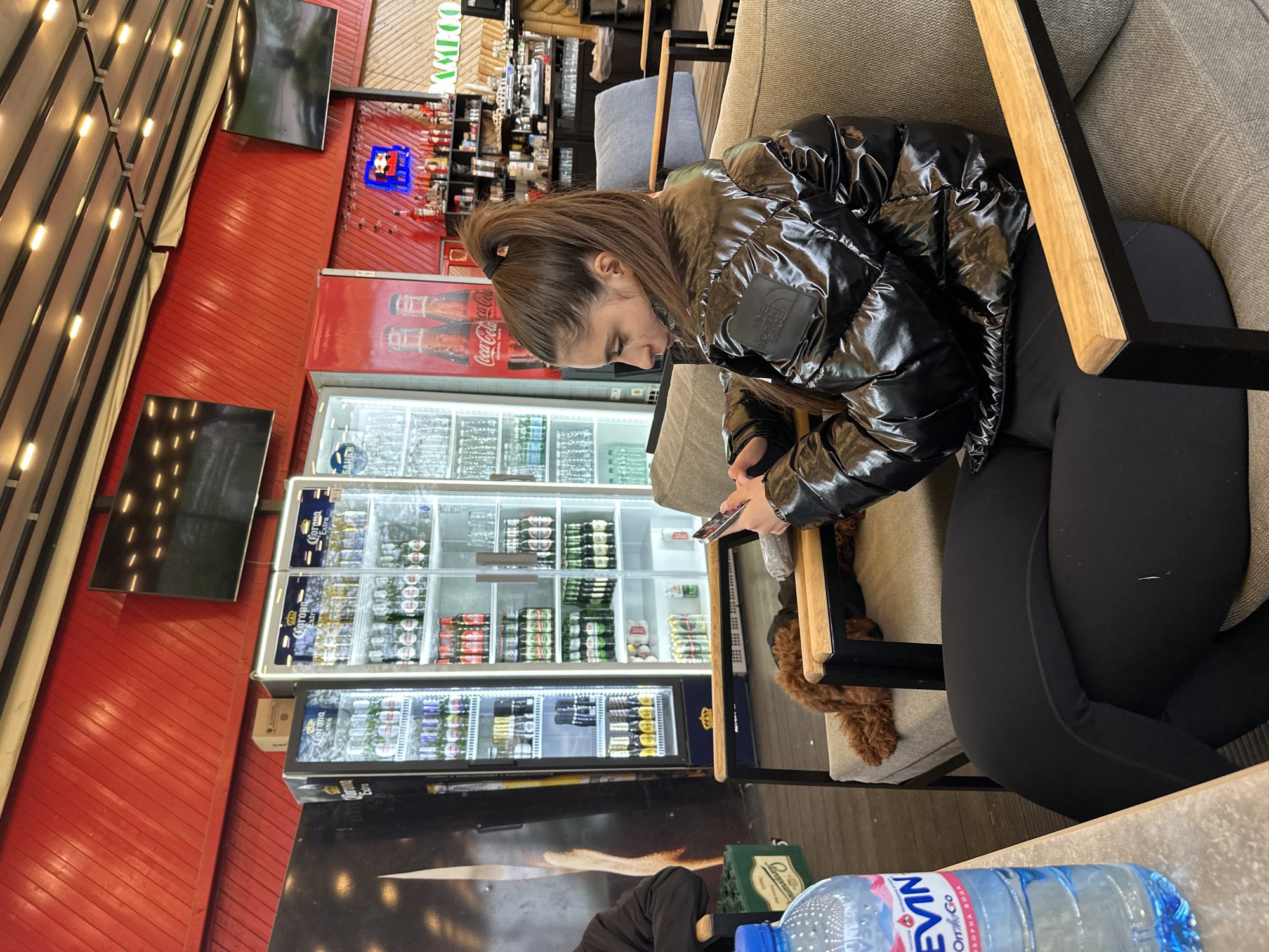 An indoor scene captures a young woman seated on a grey upholstered chair, engrossed in her smartphone. She is wearing a shiny black puffer jacket with "The North Face" logo visible on the left arm and black leggings. Her hair is pulled back in a ponytail. A small, brown, curly-haired dog is partially visible resting on the chair next to her leg.
The setting appears to be a commercial establishment like a cafe, bar, or restaurant in Plovdiv, Bulgaria. In the background, a vibrant red slatted wall runs horizontally, above which are several dark flat-screen televisions. A row of industrial-style ceiling lights is also visible. Along the wall, multiple glass-front refrigerators are stocked with various beverages, including bottles of "Corona Extra" beer and "Coca-Cola." Another red Coca-Cola branded refrigerator is also present. Shelves behind a counter display bottles and other items.
On the table in the foreground, an "EVIN On the Go" water bottle with a blue cap is prominent, next to a green crate, possibly containing beverages. Visible text on the refrigerators includes "Corona Extra," "Coca-Cola," and "БЛИКИ" (BLIKI, likely Cyrillic). A sign in the distance also displays "KYDOO" in white text against a green background. The lighting is artificial, suggesting an indoor environment at an unspecified time of day.