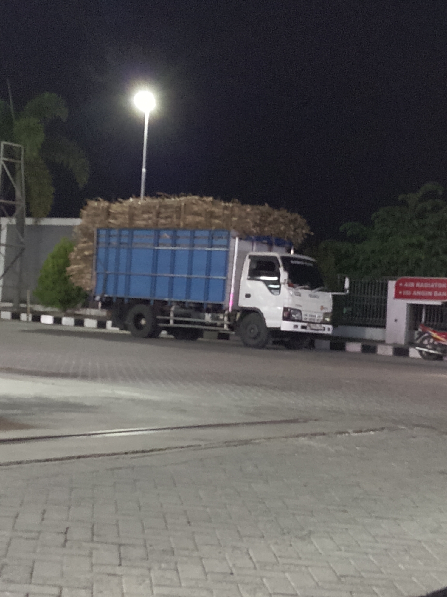 The image depicts a nighttime scene in Kasiman, Indonesia, featuring a white truck loaded with what appears to be sugarcane or similar agricultural material. The truck has a blue cargo box and is parked on a paved surface. In the background, a tall street lamp illuminates the area, and trees are visible. A metal fence with a white sign is also present. The lighting suggests the photograph was taken after dark.