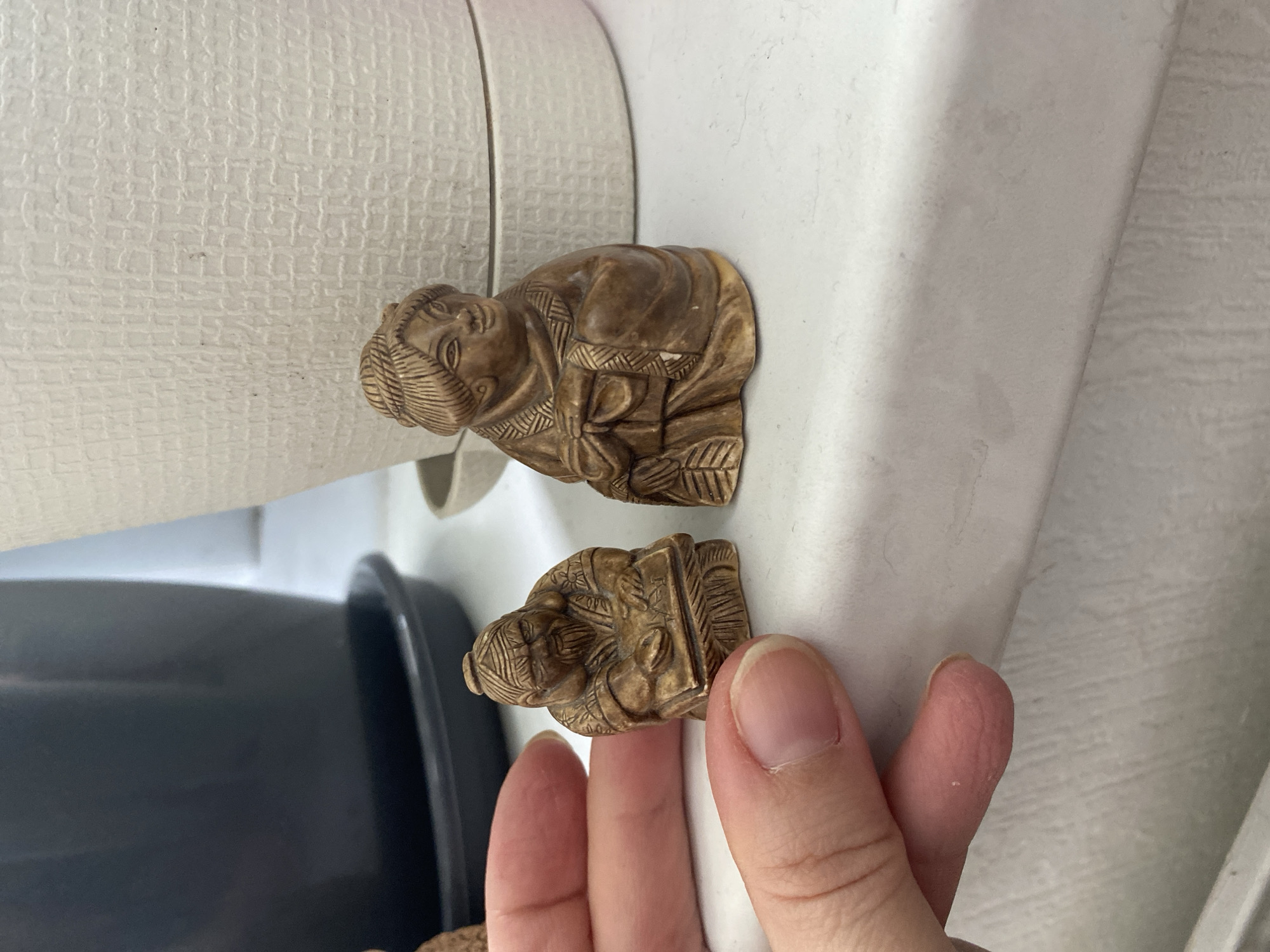 The image shows two small, carved figurines depicting human figures. They appear to be made of a light brown or tan material, possibly wood or resin. The top figurine is a detailed depiction of a person holding an object. The bottom figurine appears to have similar features. A person's fingers are partially visible in the image, near the bottom figurine. The background consists of a cream-colored, textured surface, likely a wall. A black object is visible in the background. The location of the image is not identifiable as being in Yekaterinburg, Russia, or any other specific place.