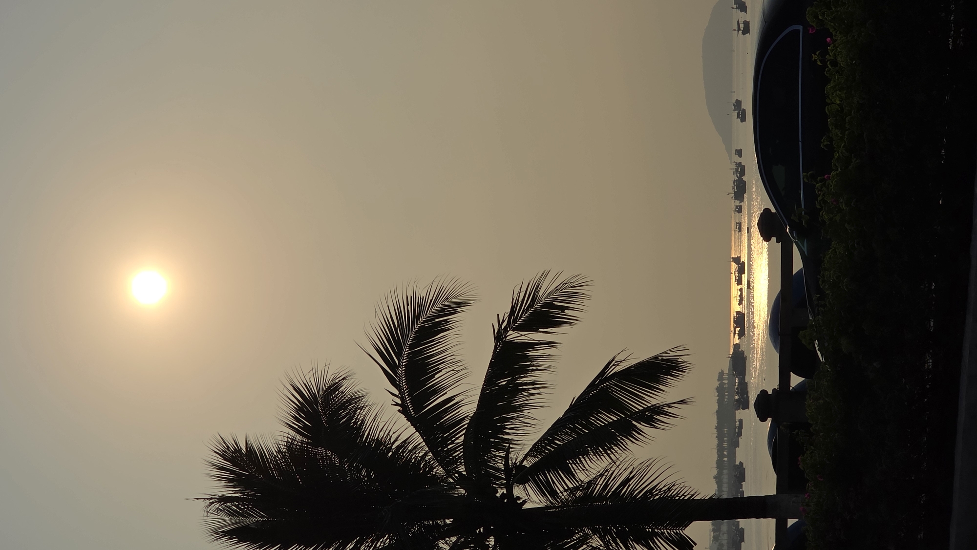 A landscape photograph captures a sunset over the city of Bắc Nha Trang, Vietnam. In the foreground, the silhouette of a coconut palm tree is featured, framing the scene. The sun is a bright, circular object positioned in the upper left of the frame. The water reflects the sunlight, creating a shimmering effect. Fishing boats dot the horizon, contributing to the coastal scenery. In the background, a row of dark objects resembling bollards are visible, along with some foliage. The sky is a gradient of light yellow and gray, indicating either sunrise or sunset. A low-lying mountain range can be seen in the distance. The image is vertically oriented.