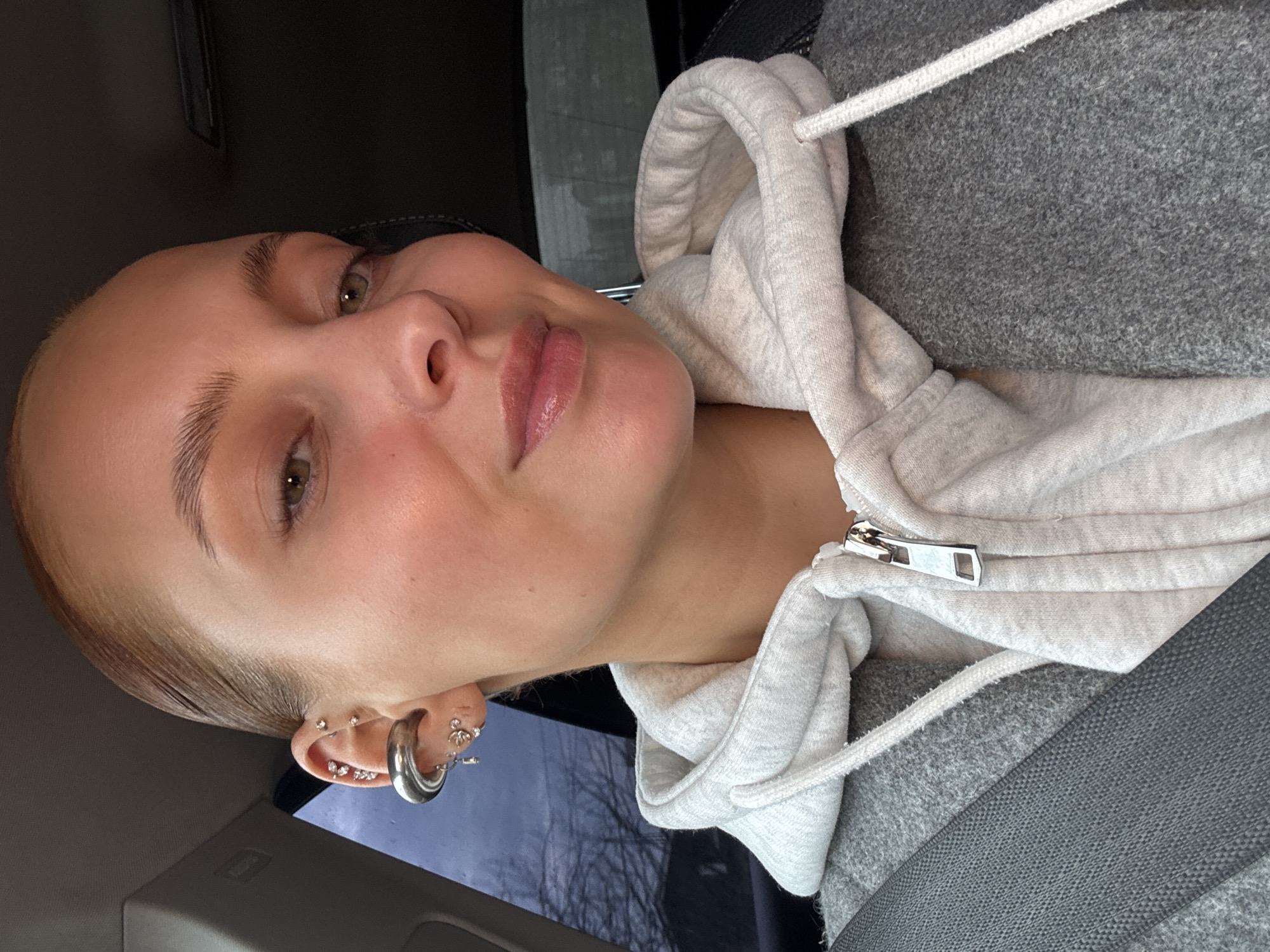 A close-up portrait shows a young person, likely female, seated inside a vehicle in Chaikovsky, Russia. The subject's head is tilted, and the image is oriented vertically, with her head on the left. She looks towards the camera with a slight, natural smile.
She has fair skin, light brown or auburn hair pulled back, and striking green/hazel eyes. Her eyebrows are well-defined, and her lips appear full and moisturized. Her left ear is visible, adorned with an array of metallic piercings, including several small studs along the cartilage, a prominent thick silver hoop in the lobe, and other smaller hoops and studs.
The subject is wearing a light grey, fleecy, zippered hoodie with a white drawstring, layered over a darker, textured grey garment. A dark grey seatbelt is visible across her chest, confirming the setting inside a car or similar vehicle. The background behind her is dark and out of focus, showing indistinct reflections that suggest outdoor scenery viewed through a window. The lighting is soft and even, indicating daytime, possibly overcast conditions. No visible text appears in the image.