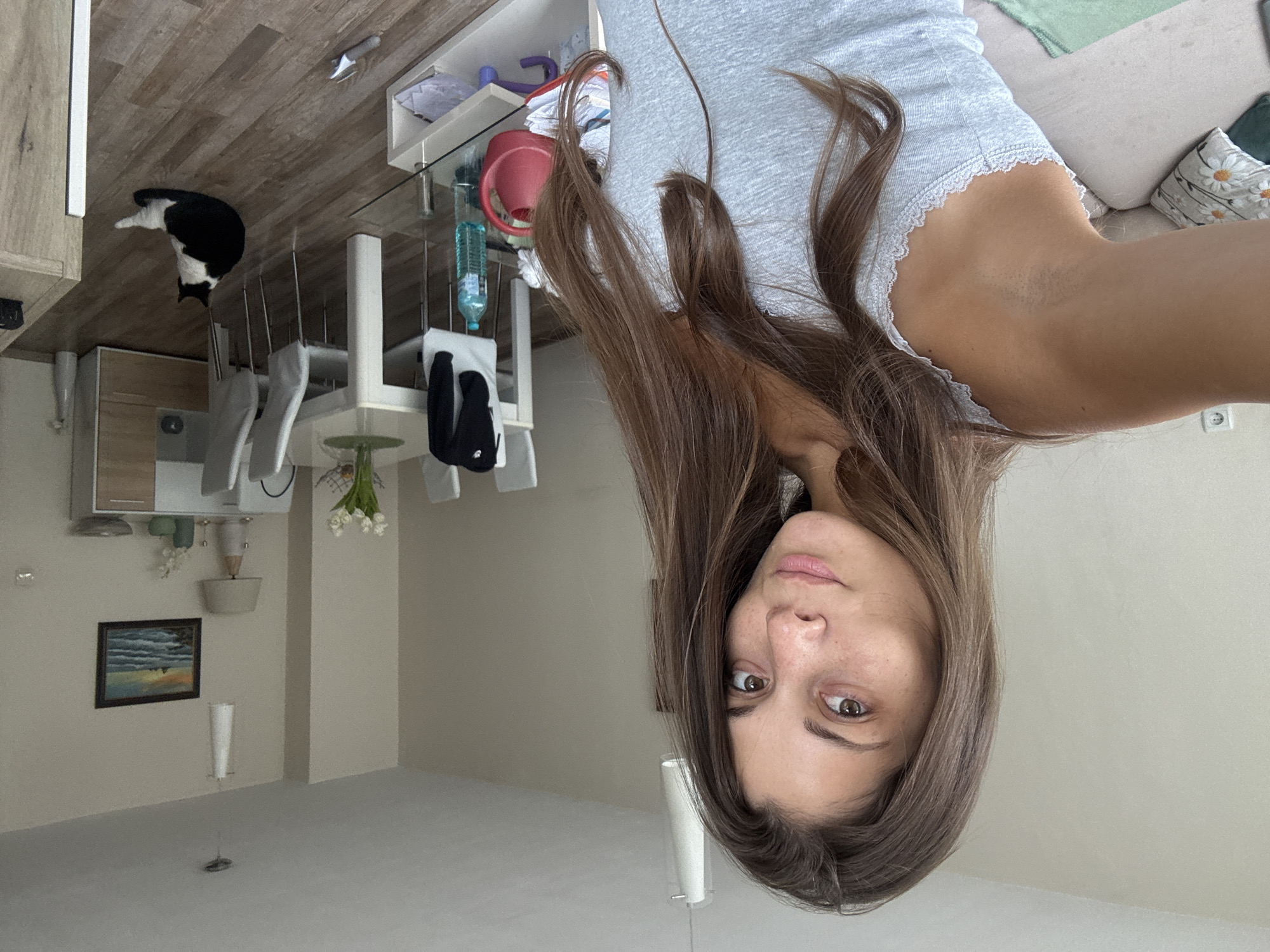 The image shows a selfie taken by a woman with long brown hair wearing a gray top with lace trim. She is indoors, in what appears to be a kitchen and living room area. The room features light-colored walls and flooring. In the background are kitchen cabinets, a dining table with chairs, a shelf with various items including a red watering can and cleaning supplies, a black and white cat, and a painting on the wall. In the living room area, there is a couch with pillows. There is no indication that the image was taken in Burgas, Bulgaria.