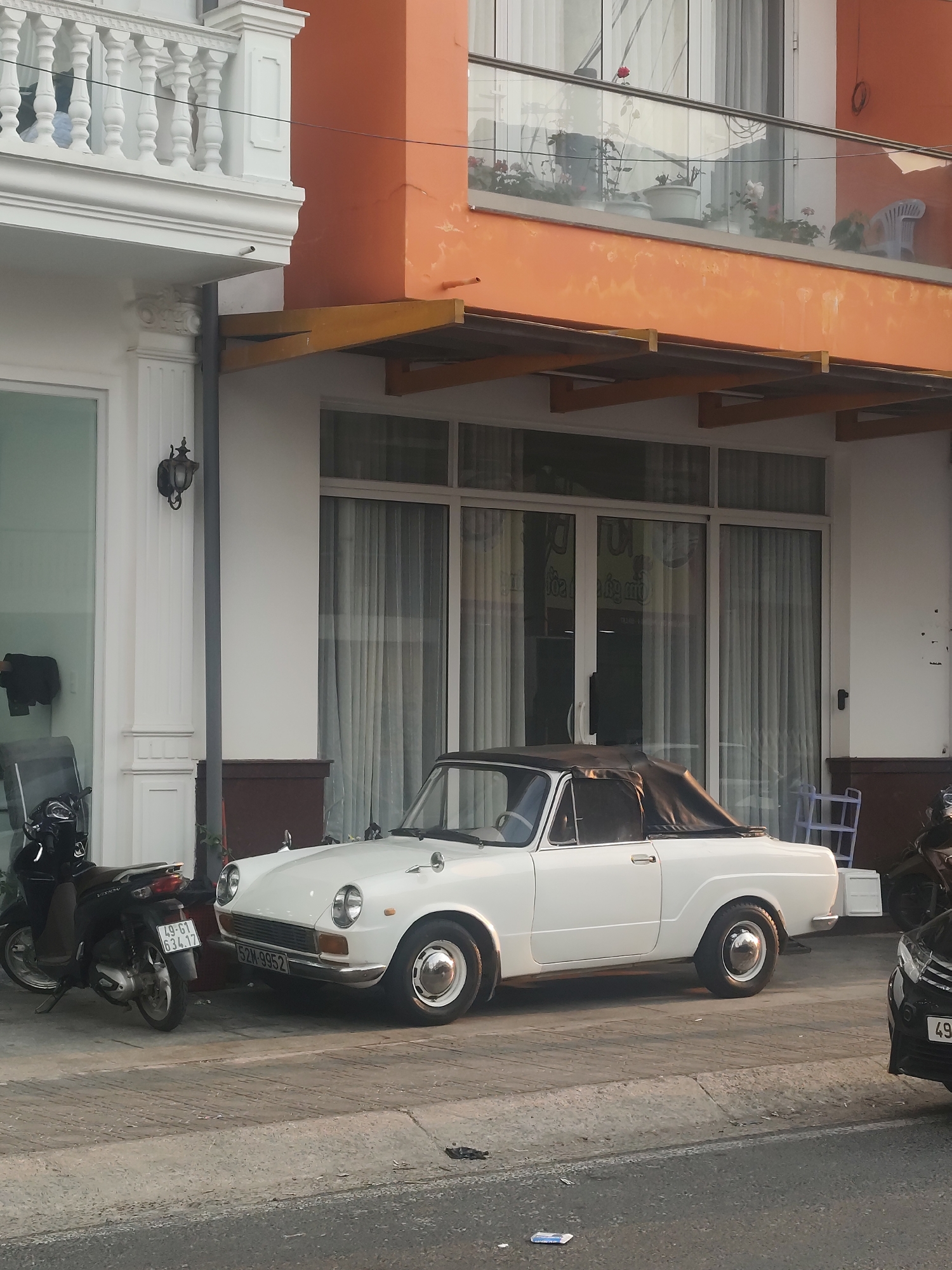 The image captures a street scene in Da Lat, Vietnam. A vintage white convertible car with a black soft top is parked along the sidewalk. To the left of the car is a black scooter, and to the right, the front of a black sedan is visible. The car's license plate reads "52M-9952," while the scooter's plate shows "49-G1 634.17."
Behind the vehicles, a building with a white facade and an orange overhanging structure is present. The building features large glass windows with translucent curtains inside. A balcony above the orange overhang displays potted plants and a white plastic chair. The overall environment appears urban, with elements of classic European architecture mixed with Southeast Asian vehicles and building styles.