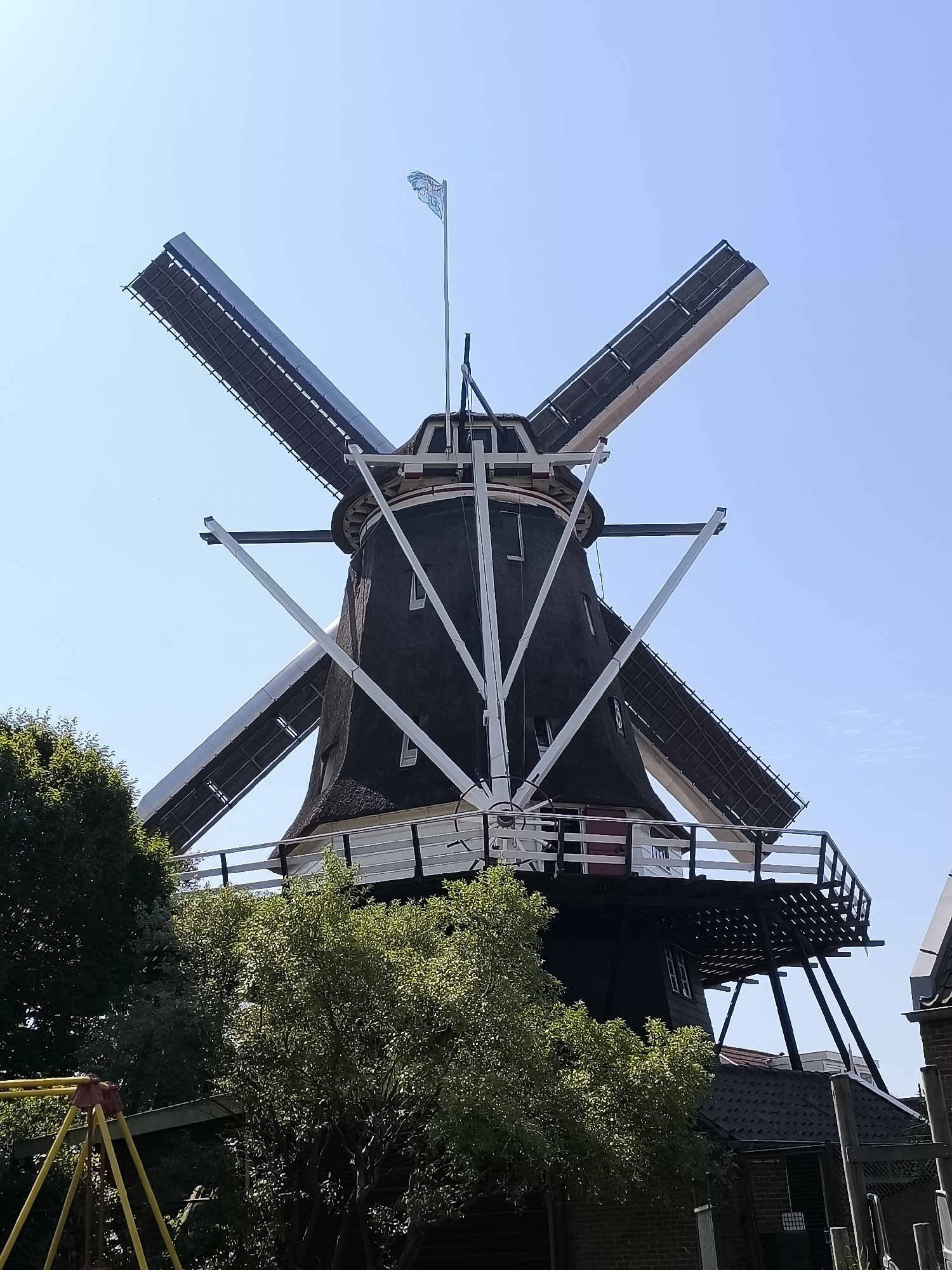 The image shows a close-up, low-angle view of a traditional Dutch windmill under a clear blue sky. The windmill has a dark, thatched roof and four large sails, each with a white frame and dark fabric panels. White structural elements are visible supporting the sails and the cap of the windmill. A small Dutch flag, blowing in the wind, is mounted on a pole at the very top of the windmill. The lower part of the windmill has a black exterior with a white railing around a platform. Green trees with lush foliage are visible in the foreground, partially obscuring the base of the windmill. A yellow structure, possibly part of a playground, is visible on the left side of the image. The windmill is located in the city of Alkmaar, Netherlands.