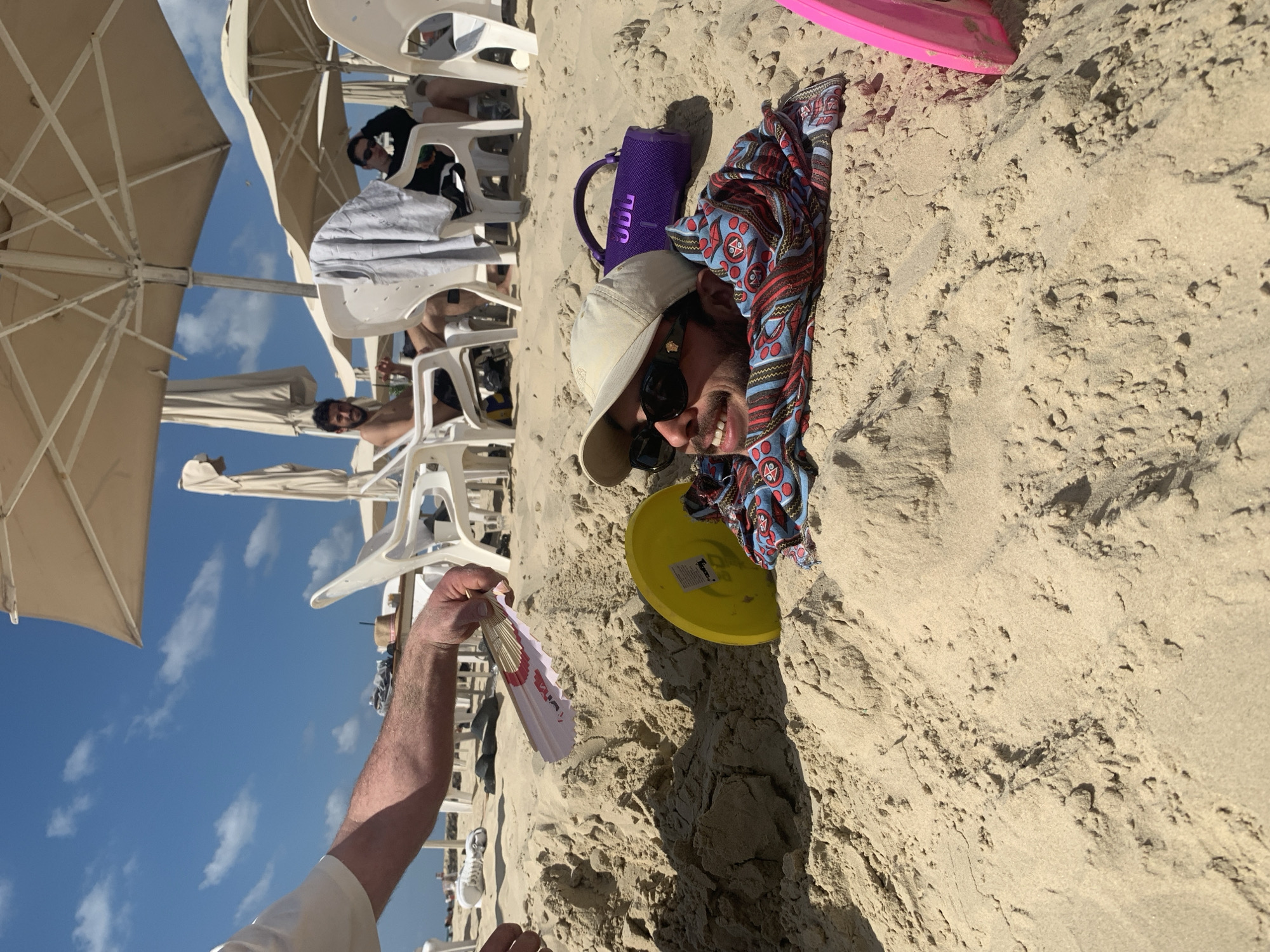 This image captures a vibrant and playful scene at a sandy beach, likely in Tel-Aviv, Israel, under a bright blue sky with scattered clouds.
In the mid-ground, a man with a beard, wearing sunglasses, a beige cap, and a colorful patterned shirt, is partially buried in the sand, smiling broadly at the camera. Beside his head rests a purple JBL brand speaker and a yellow frisbee, which has "Tuggy" visible on its surface. To his right, a pink plastic spade is half-buried in the sand.
In the foreground, an arm extends from the left side of the frame, holding a white and pink fan, suggesting someone is fanning themselves or another person in the warm weather.
The background reveals a typical beach setup with numerous white plastic chairs and large beige umbrellas providing shade, under which other people are seen relaxing. The overall impression is one of a cheerful and leisurely day at the beach, with clear skies and ample sunlight.
