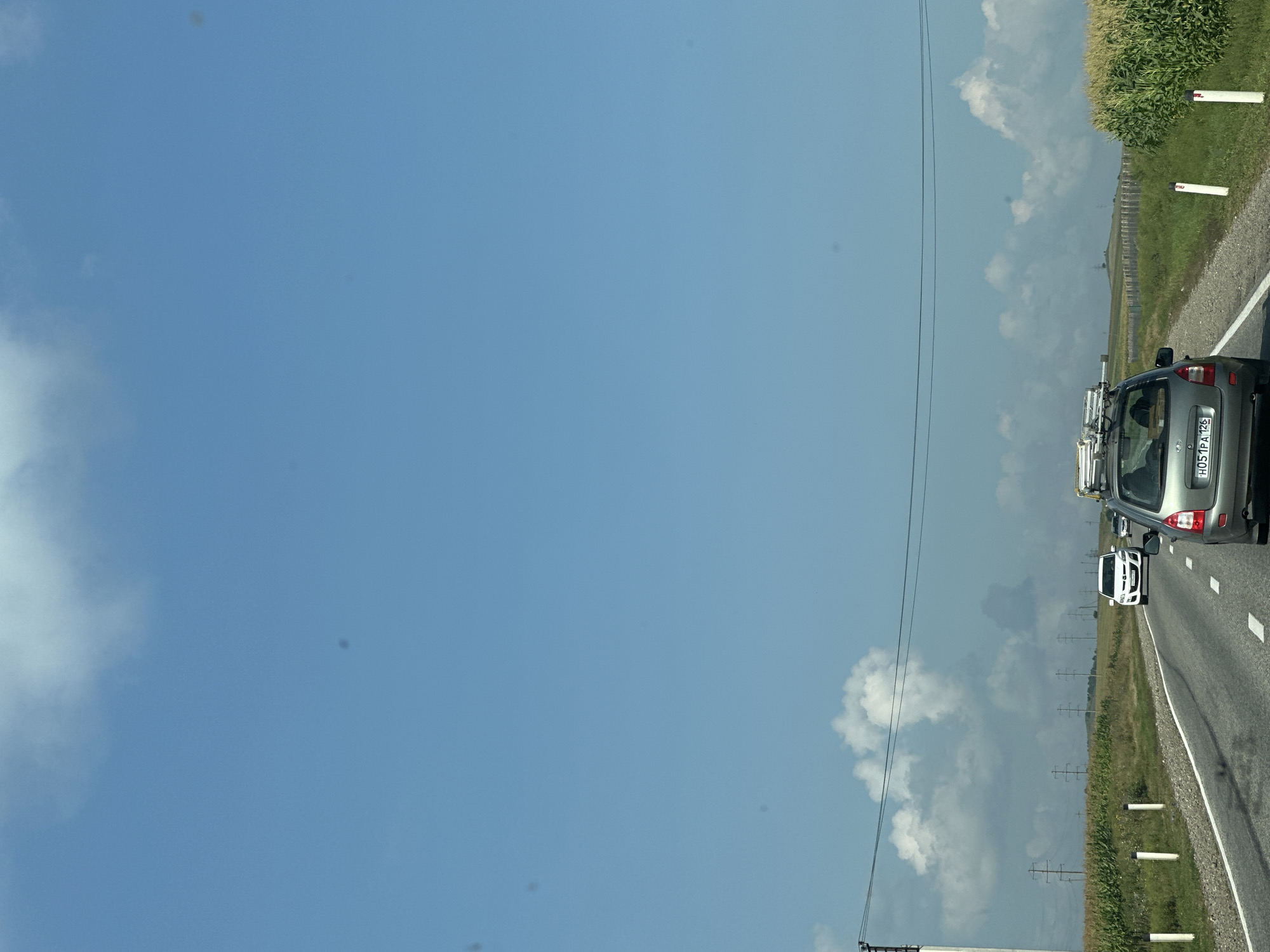 The image depicts a road in Kardonikskaya, Russia. A gray car is traveling on the asphalt road, with a white car in front of it and a cornfield on the right side of the road. The sky is blue with white clouds, and there are power lines and utility poles visible in the background.