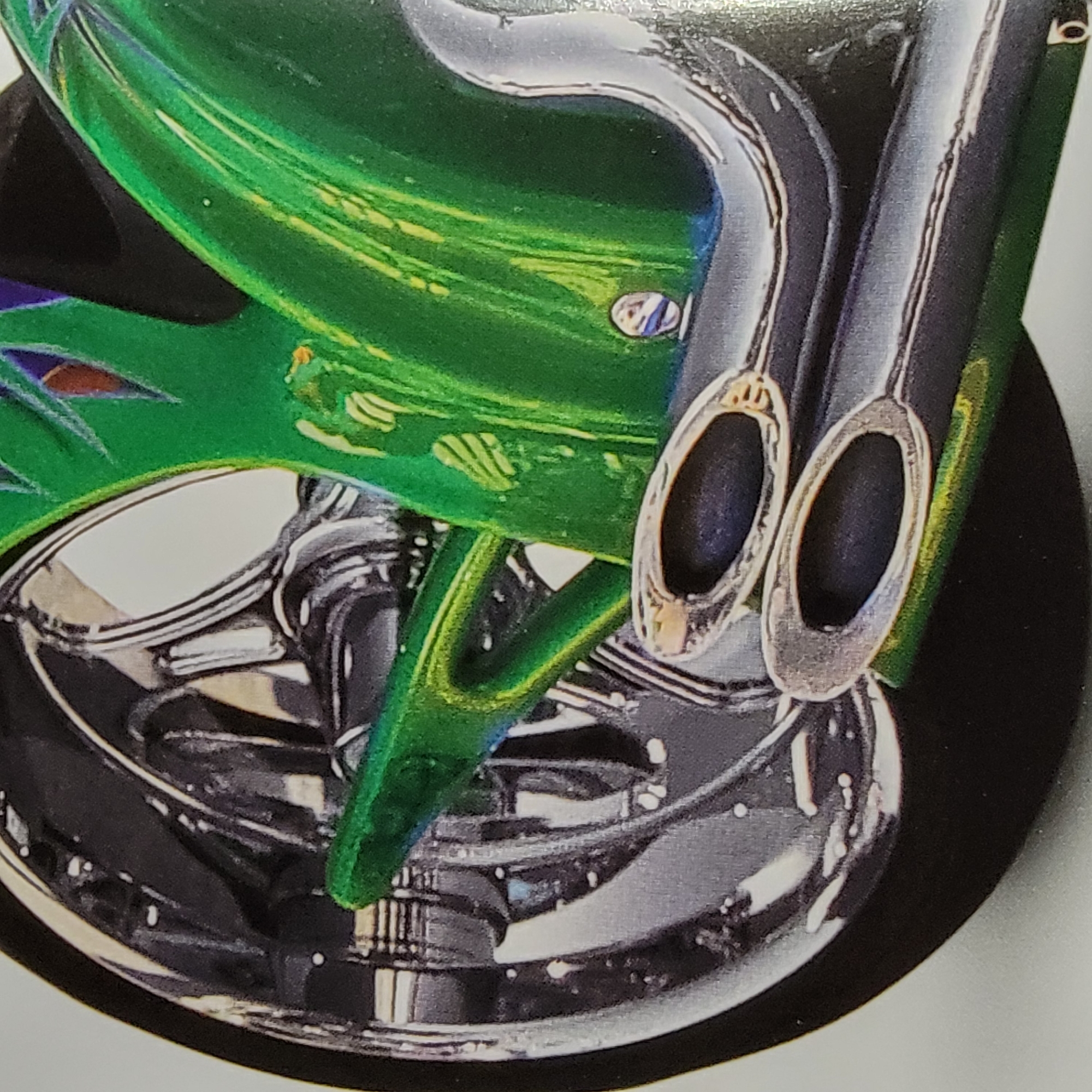 The image shows a close-up of a motorcycle's rear wheel, exhaust pipes, and part of the green bodywork. The wheel is chrome with a black tire. Two silver exhaust pipes with chrome tips are visible in the foreground. The bodywork is a vibrant green with yellow and blue flame-like graphics. The background is white. The image is a photograph of a motorcycle, likely taken in a studio or outdoor setting, but there are no identifiable landmarks to suggest a specific city or location.