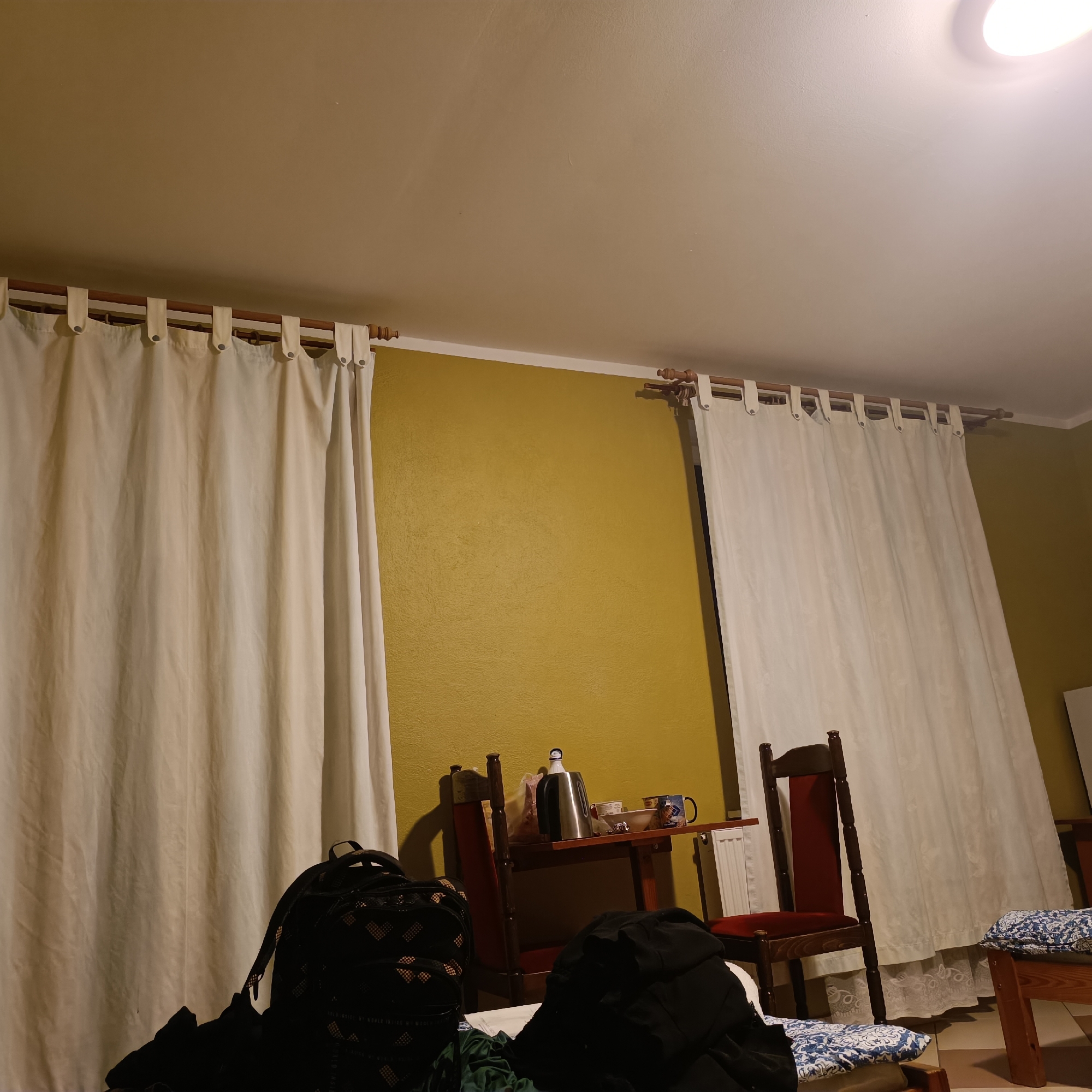 The image shows an indoor scene, likely a room in a guesthouse or hotel in Mielno, Poland. The room has yellow walls and is furnished with white curtains, a small wooden table with chairs, and some luggage. On the table, there is a silver kettle, a bowl, and a mug. A white radiator is visible on the right side of the image. There are two wooden chairs with red upholstery. Luggage, including a black backpack with a heart pattern, is placed on the floor. The lighting suggests it is evening or dimly lit.