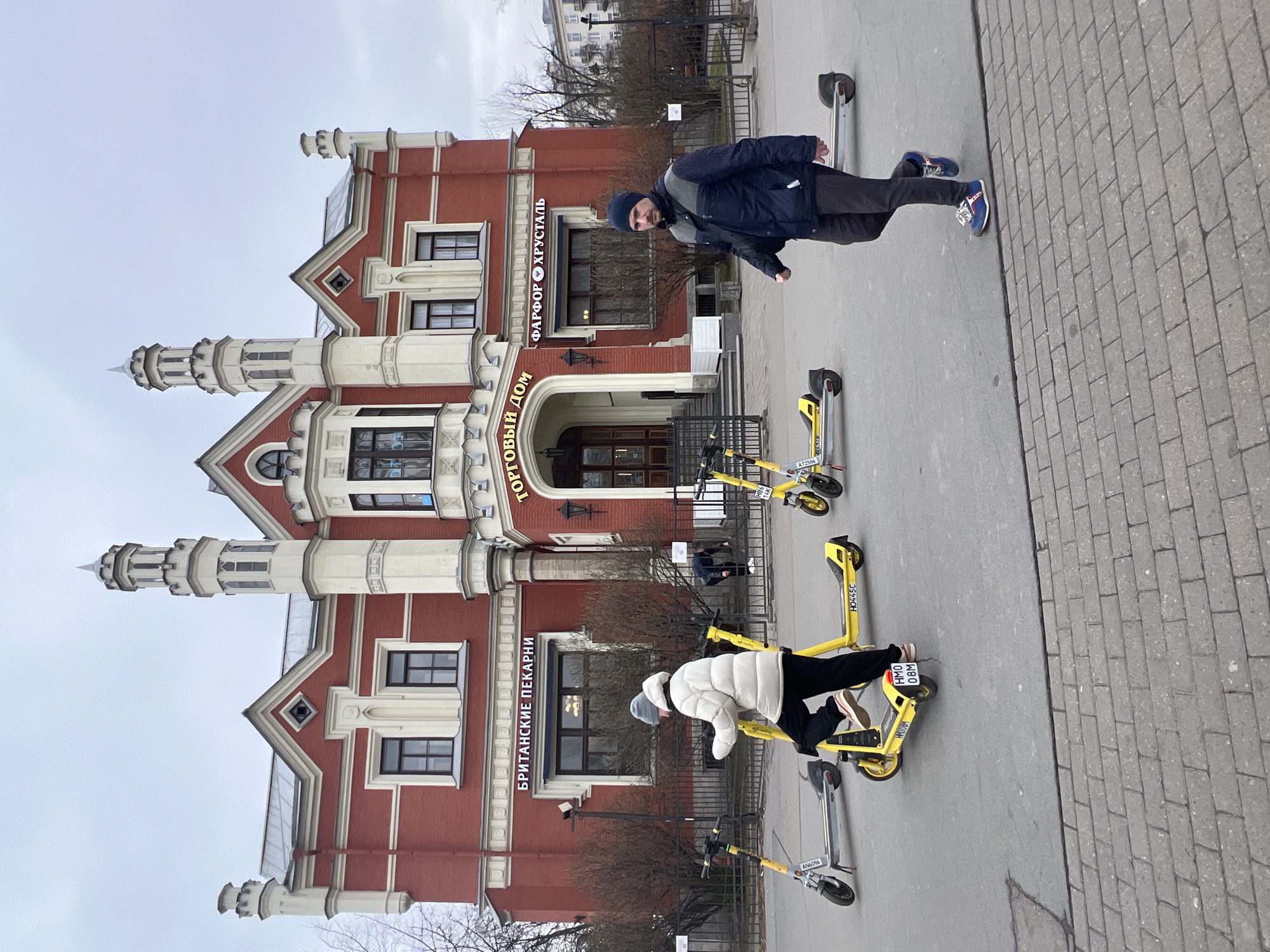 The image shows the facade of the building at Nevsky Prospekt 47 in Saint Petersburg, Russia. The building is brick-red with white architectural details and has two decorative towers. Signage in Russian is visible on the front of the building. Two people are standing on the sidewalk in front of the building, posing next to yellow rental scooters. The sidewalk is paved with grey brick. The sky is overcast.