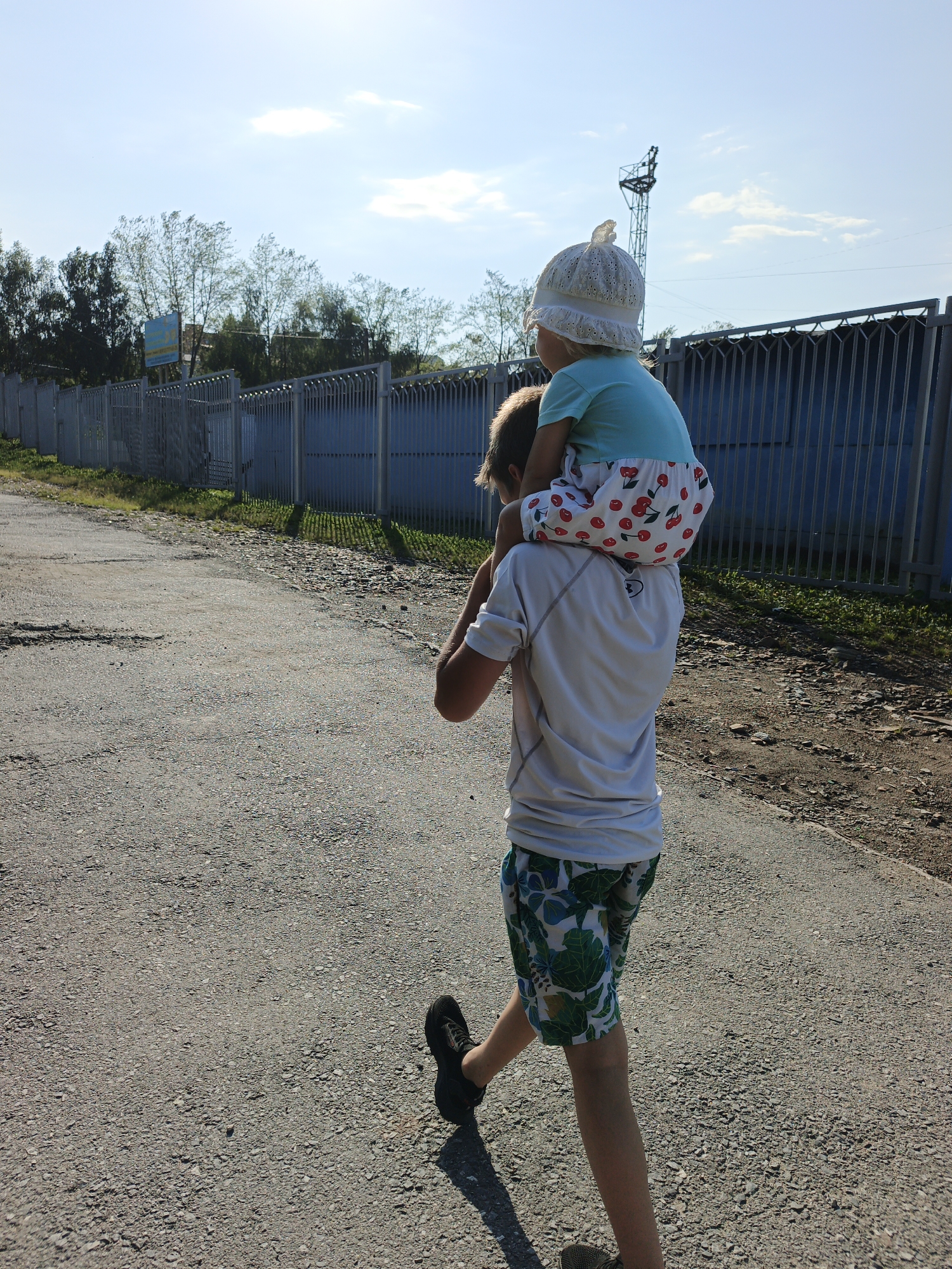 A boy carrying a young girl on his shoulders walks along an asphalt road in Zlatoust, Russia. The boy, wearing a white t-shirt and patterned shorts, is viewed from behind as he walks away from the camera. The girl, also facing away, wears a light blue t-shirt and a white hat, with a white skirt patterned with red cherries. The road is flanked by a gravel shoulder and a tall metal fence, with trees and a utility tower visible in the background under a bright sky. The scene suggests a casual outdoor activity on a sunny day.