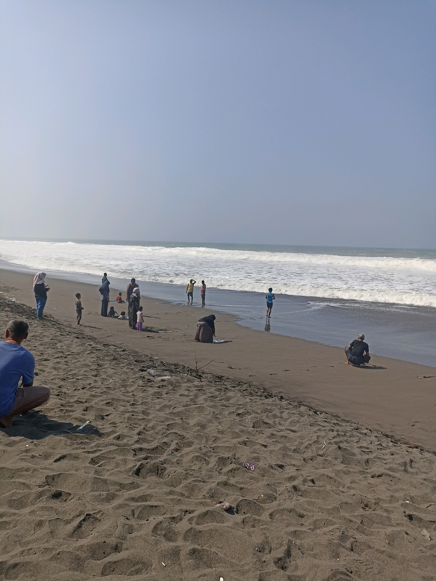 In the city of Kenoyajaya, Indonesia, a group of people are gathered on a sandy beach near the ocean. The scene is captured under a clear, light blue sky with hazy conditions near the horizon. The ocean features large, white waves breaking on the shore.
In the foreground, on the dark sand, a person wearing a blue t-shirt and light-colored shorts is sitting and facing away from the camera, looking towards the ocean. To the right, another person is crouching on the sand, also facing the ocean, wearing a dark cap and patterned shorts.
Further back, along the water's edge, several individuals are visible. A woman wearing a hijab and a blue top stands near the left side of the frame, facing left. Beside her, a child in a grey outfit stands. A group of people are congregated near the center, including a woman in a dark hijab and long skirt, a child in red, and other individuals in the background. Two children are playing near the water's edge. A man in a yellow shirt is standing further down the beach.
The sand is dark and textured, with some scattered debris, including plastic items, visible on the surface. The overall impression is of a casual gathering at the beach on a bright day.