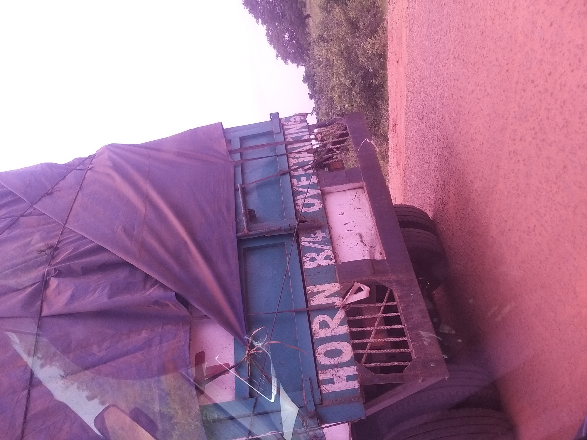 The image shows the rear of a blue truck covered with a purple tarp, parked on a dirt road. The truck has "HORN B/4 OVERTAKING" painted on its side in white letters. There are visible tires on the truck and a metal grate at the back. The surrounding environment consists of dry vegetation and trees, with a hazy sky in the background. The scene appears to be in Dukku, Nigeria, based on the provided location information, likely in a rural or semi-urban area given the dirt road and natural surroundings.