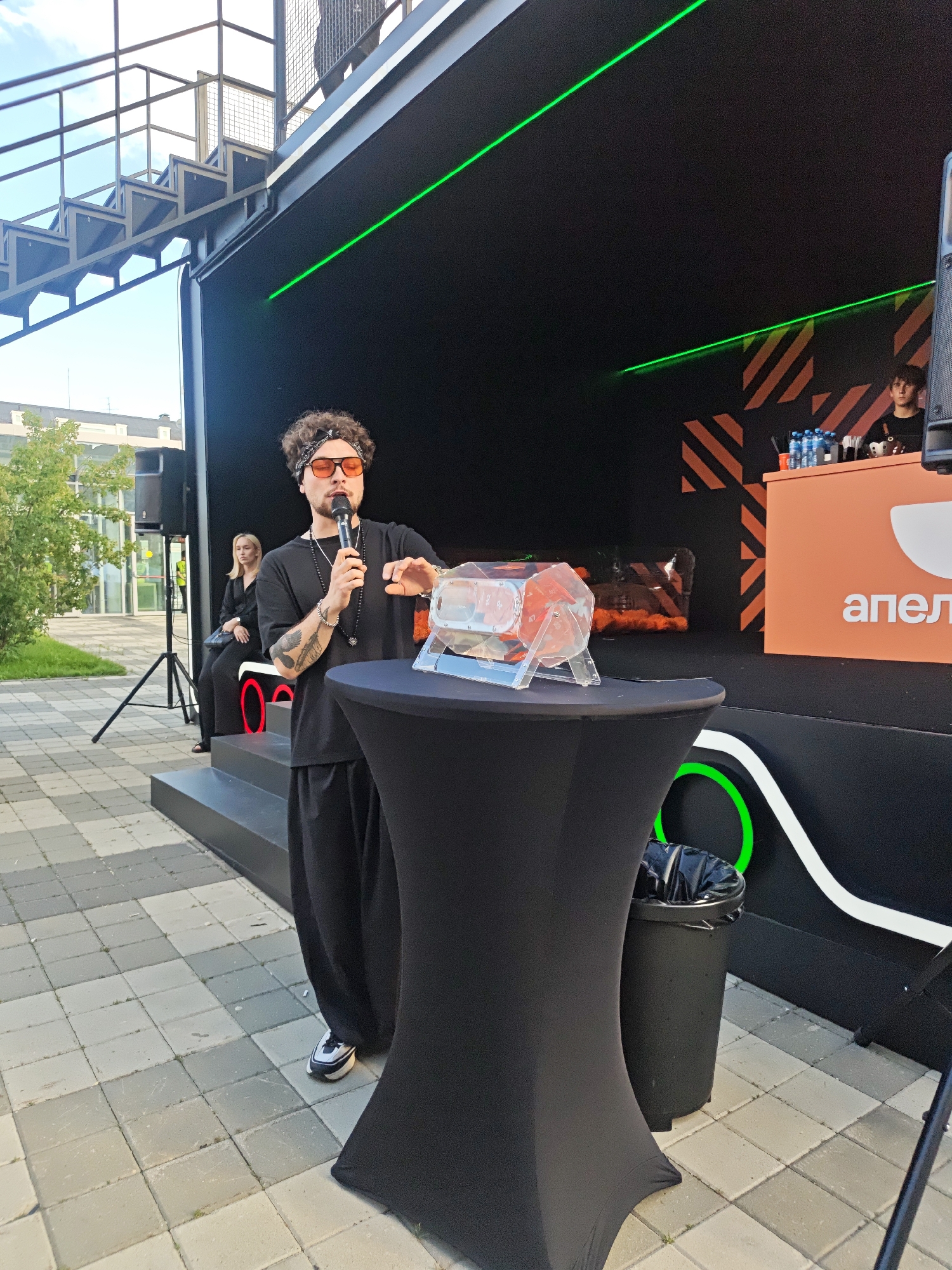 A man with curly hair, sunglasses, and a bandana is speaking into a microphone at an outdoor event. He is standing behind a black tablecloth-covered table on which a clear container with red and clear plastic elements is placed. He is wearing a black t-shirt, black pants, and white and black sneakers. Behind him, a woman in a black outfit is seated on steps. The background features a black structure with green neon accents, a metallic staircase, and a white and orange structure with text. A trash can is to the right of the man. The ground is paved with square tiles. The scene is likely taking place in a public space in Yekaterinburg, Russia, given the visible Cyrillic text "апел" on the orange structure, which translates to "apple".