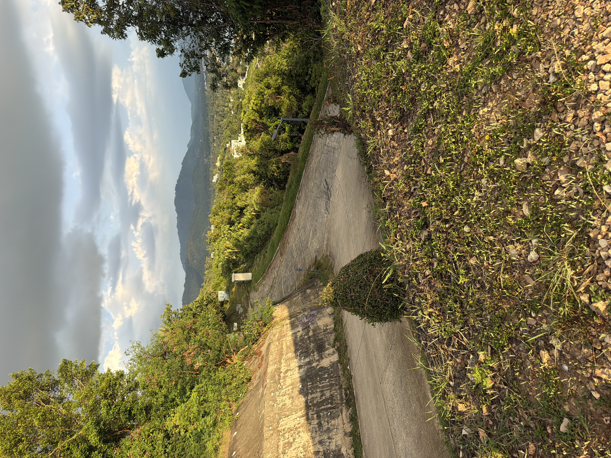 The image depicts a paved road winding uphill through a lush, green landscape under a dramatic sky with both dark clouds and patches of sunlight. The road is bordered by a concrete retaining wall on the left, showing signs of age and some graffiti, and dense vegetation on the right. Further up the road, a street lamp is visible, and a small, rectangular structure stands on the side. Beyond the immediate foreground, the scene opens up to a vista of rolling hills covered in trees, with scattered buildings visible in the distance, suggesting a residential area within a tropical or subtropical environment. The sunlight casts long shadows, indicating it might be late afternoon or early morning. The overall impression is one of a rural or semi-rural setting, possibly on the outskirts of a town or city, with natural beauty contrasted by man-made infrastructure.
