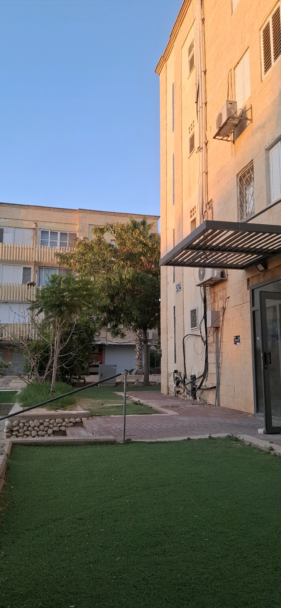 The image depicts a residential building in Yerocham, Israel, bathed in the warm glow of late afternoon sun. The building is constructed of light-colored stone blocks, with multiple windows, some featuring white shutters. An air conditioning unit is mounted on the exterior wall, with exposed electrical conduits and pipes running vertically. Above the entrance to the building, a dark metal awning casts a shadow. The building number "534" is visible on the facade. To the left, another building is partially visible, with a balcony featuring vertical white railings and windows. A small, green lawn area with artificial turf occupies the foreground. Adjacent to the lawn is a paved walkway made of brick pavers. A low retaining wall constructed with rounded stones borders a raised garden bed with sparse green vegetation. Trees with dense foliage are present in the midground, casting shadows. The sky is clear and blue. No people are present in the scene.