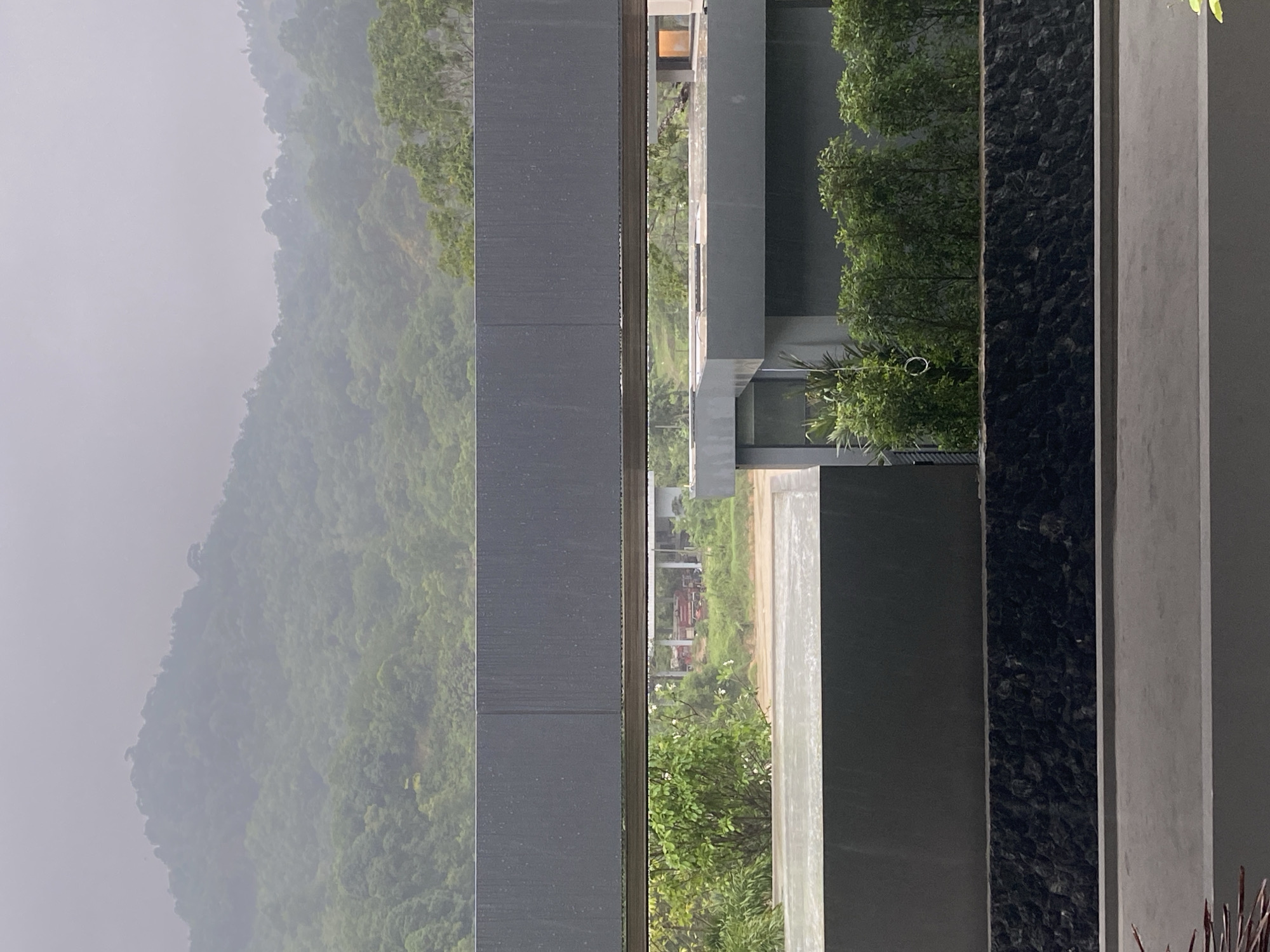 The image shows a view of a green, mountainous landscape through a series of vertical architectural elements. The location is Choeng Thale, Thailand. The architectural elements consist of gray and black columns. Through the gaps between these columns, a lush, green mountain covered in mist is visible. There are some buildings and vegetation in the foreground. The weather appears overcast, contributing to the misty atmosphere.