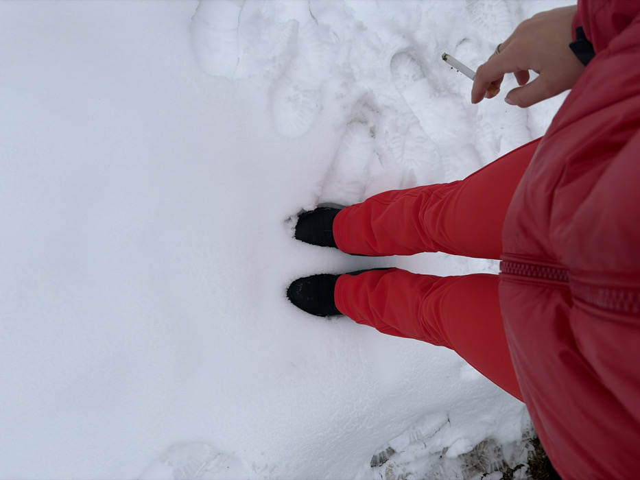 A first-person, downward perspective captures a person standing in fresh snow in La Clusaz, France. The individual wears red snow pants and black winter boots, with a portion of a red puffer jacket visible at the bottom of the frame. The left hand, adorned with a gold-colored ring on the ring finger, holds a lit cigarette between the index and middle fingers, displaying an active ember and a small amount of ash. The ground is covered in undisturbed white snow, with several depressions and faint foot impressions visible to the right of the person's feet. Patches of darker ground or ice are partially visible through the snow on the far left.