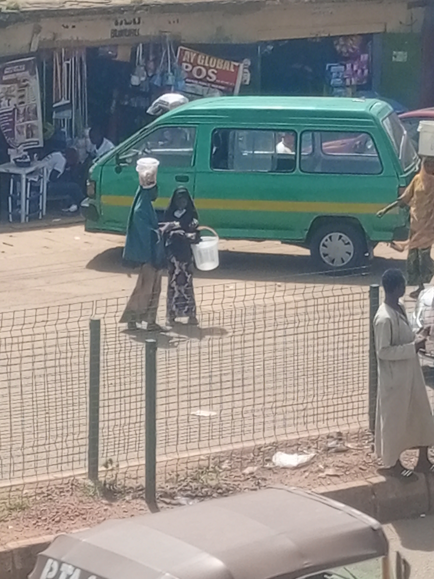 This media file depicts a street scene in Bukuru, Nigeria. The central focus is a green mini-bus with a yellow stripe, parked on a paved road. In front of the bus, two individuals are visible. One, dressed in a dark headscarf and patterned attire, carries a white bucket. Beside them, another person, wearing a blue headscarf and a flowing robe, holds a white bucket on their head.
The background reveals a row of shops with open fronts, displaying various goods. A sign above one of the shops reads "AY GLOBAL POS". Several objects, including what appear to be brooms, are hanging within the shop entrances. Other people are also present in the scene, some seated or standing near the shops, and another individual in a light-colored robe stands near a wire fence.
The lighting suggests it is daytime, with bright sunlight casting shadows. The weather appears to be clear. The ground is dusty and uneven, typical of a street in a developing urban area. The overall atmosphere seems to be one of everyday commerce and transit.